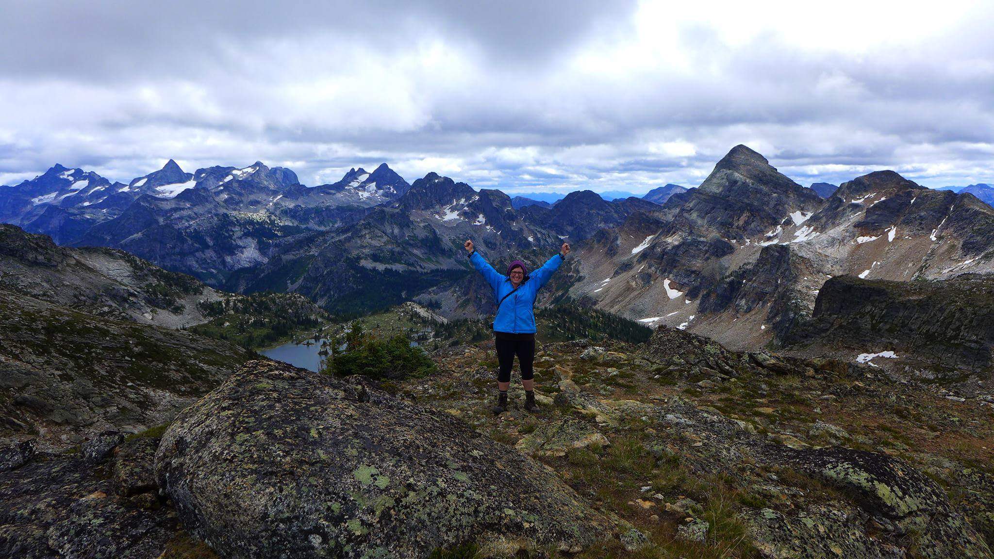 A hiker in a blue jacket stands on rocky ground with arms raised, surrounded by dramatic, jagged mountain peaks, patches of snow, and alpine lakes beneath a cloudy sky.