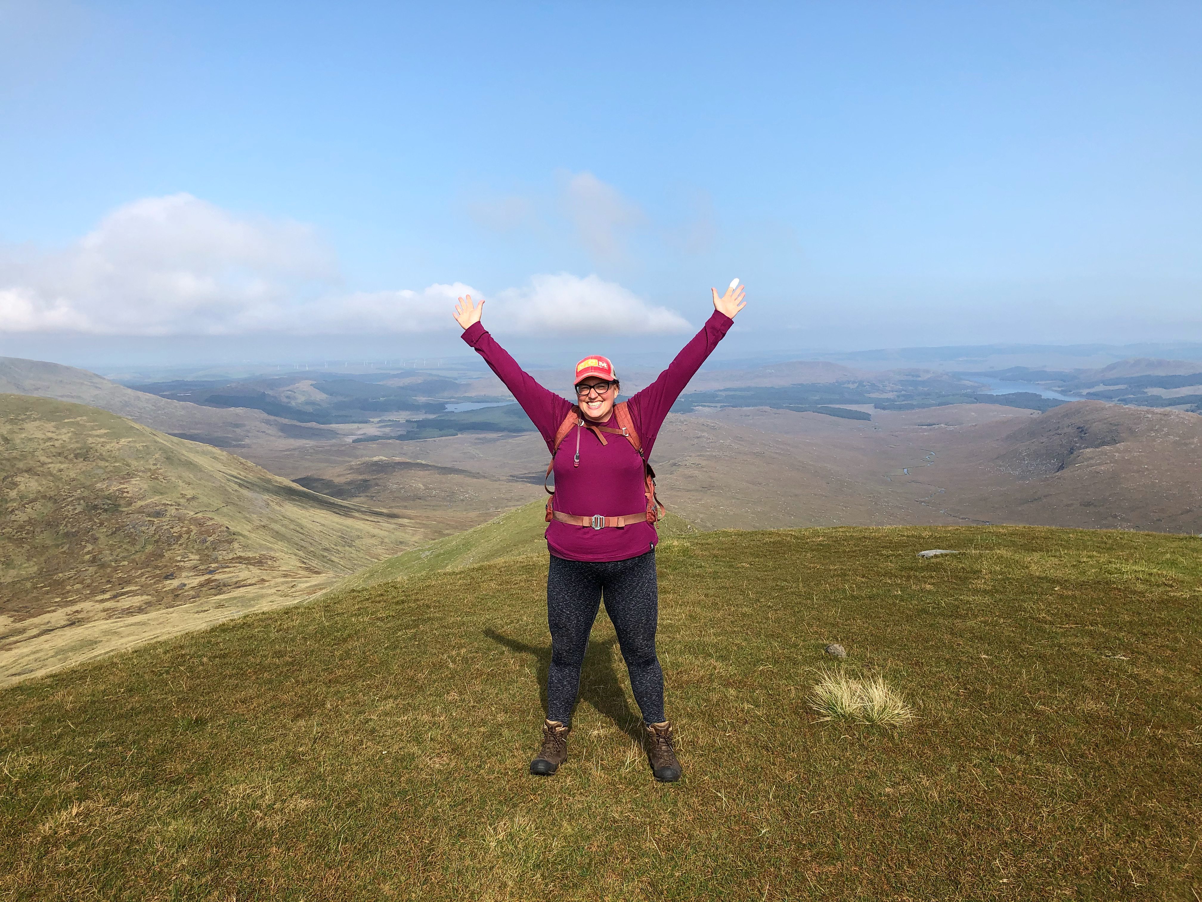 A hiker stands on a grassy hilltop with arms raised in celebration, wearing a purple top, backpack, and cap, with wide open hills, valleys, and a blue sky with light clouds in the background.
