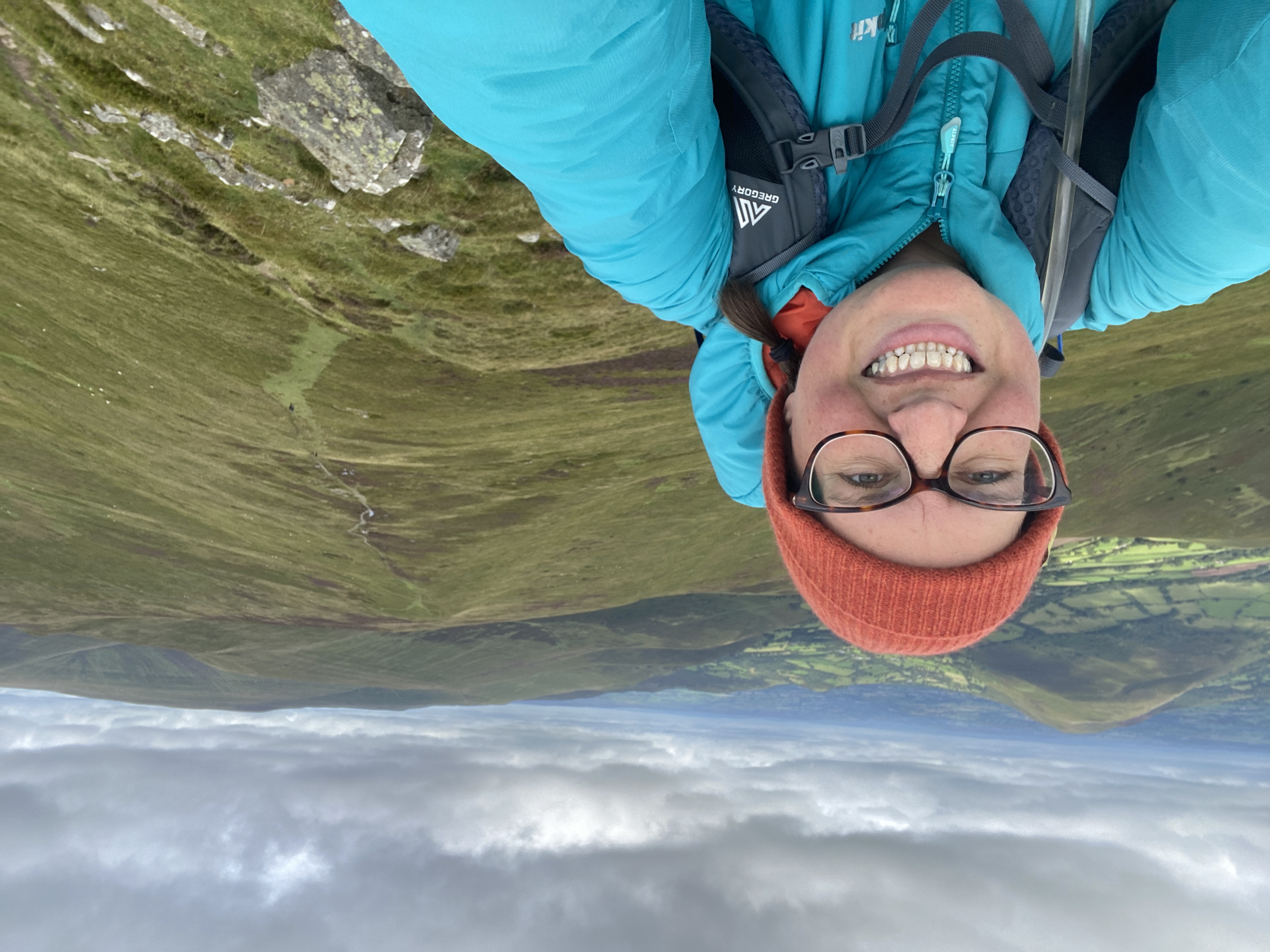 A smiling hiker on Offa’s Dyke National Trail wearing glasses, an orange knit hat, and a turquoise jacket takes a selfie on a grassy mountain ridge, with rolling green hills and a winding path stretching into the distance under a cloudy sky.