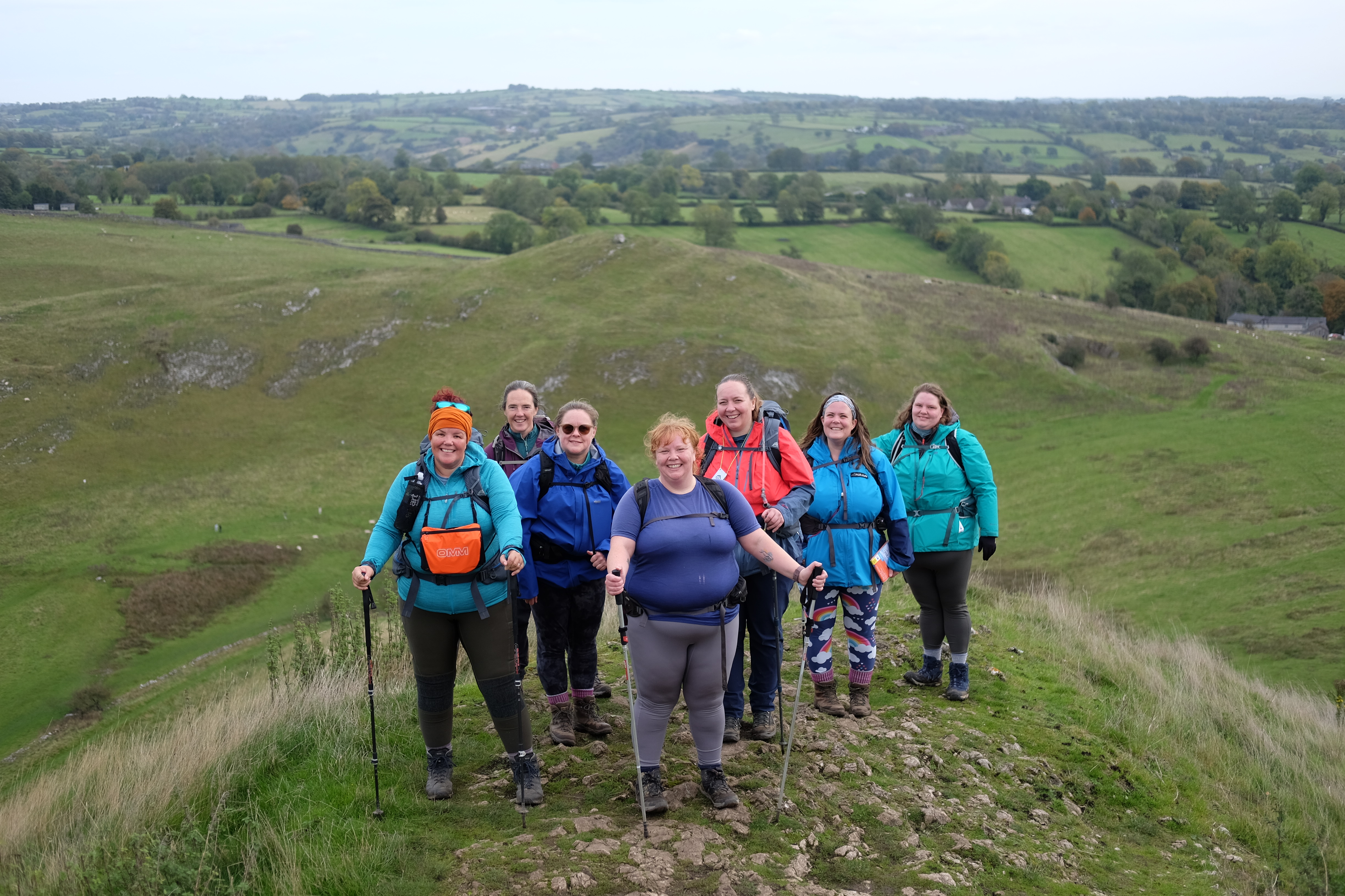 A group of eight hikers wearing colourful outdoor jackets and backpacks stand together on a grassy hillside path, smiling at the camera, with rolling green countryside and fields stretching out behind them.