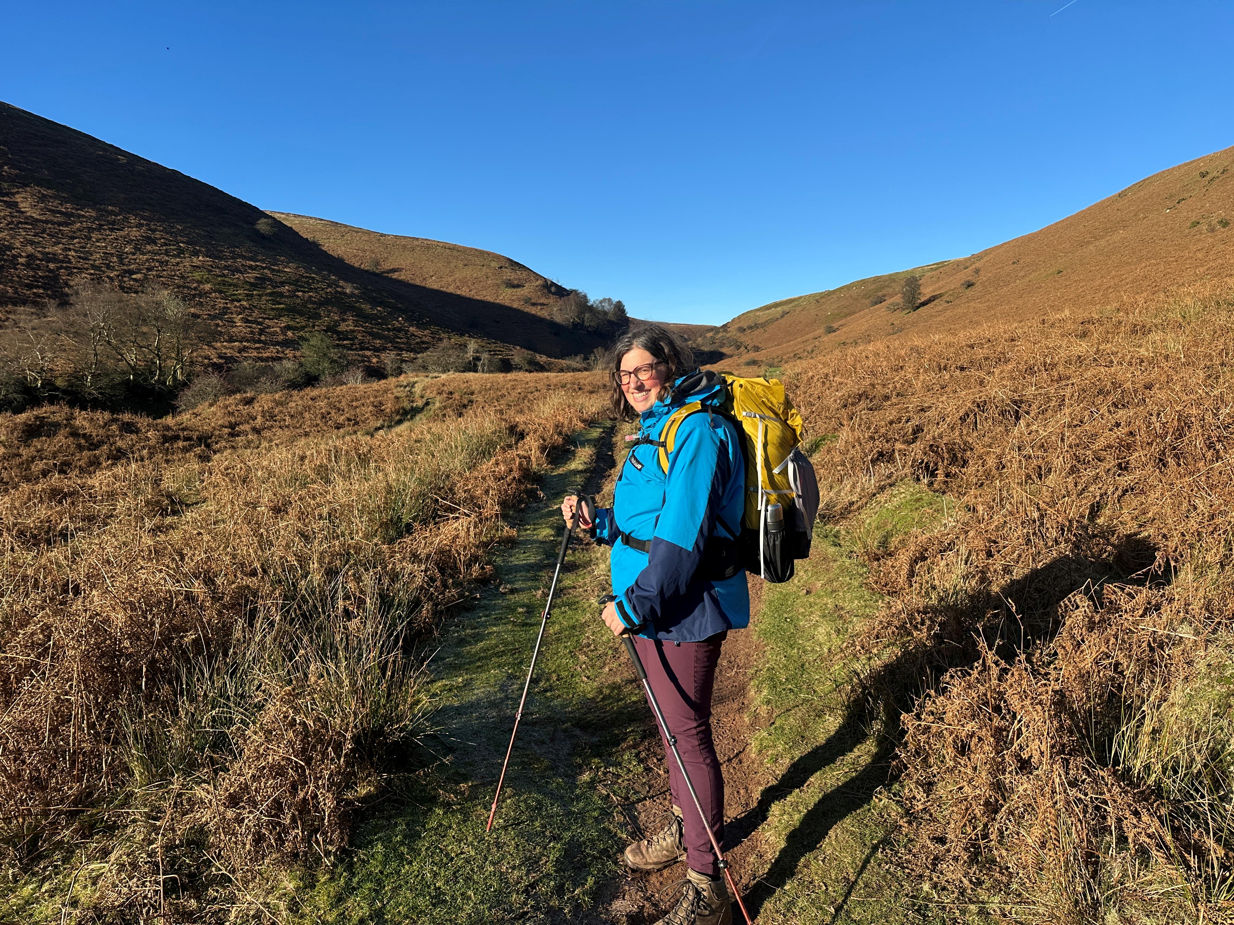 A hiker wearing a blue jacket and carrying a yellow backpack stands on a narrow grassy path between brown, bracken-covered hills. They are holding walking poles and smiling, with rolling hills and a clear blue sky in the background.”