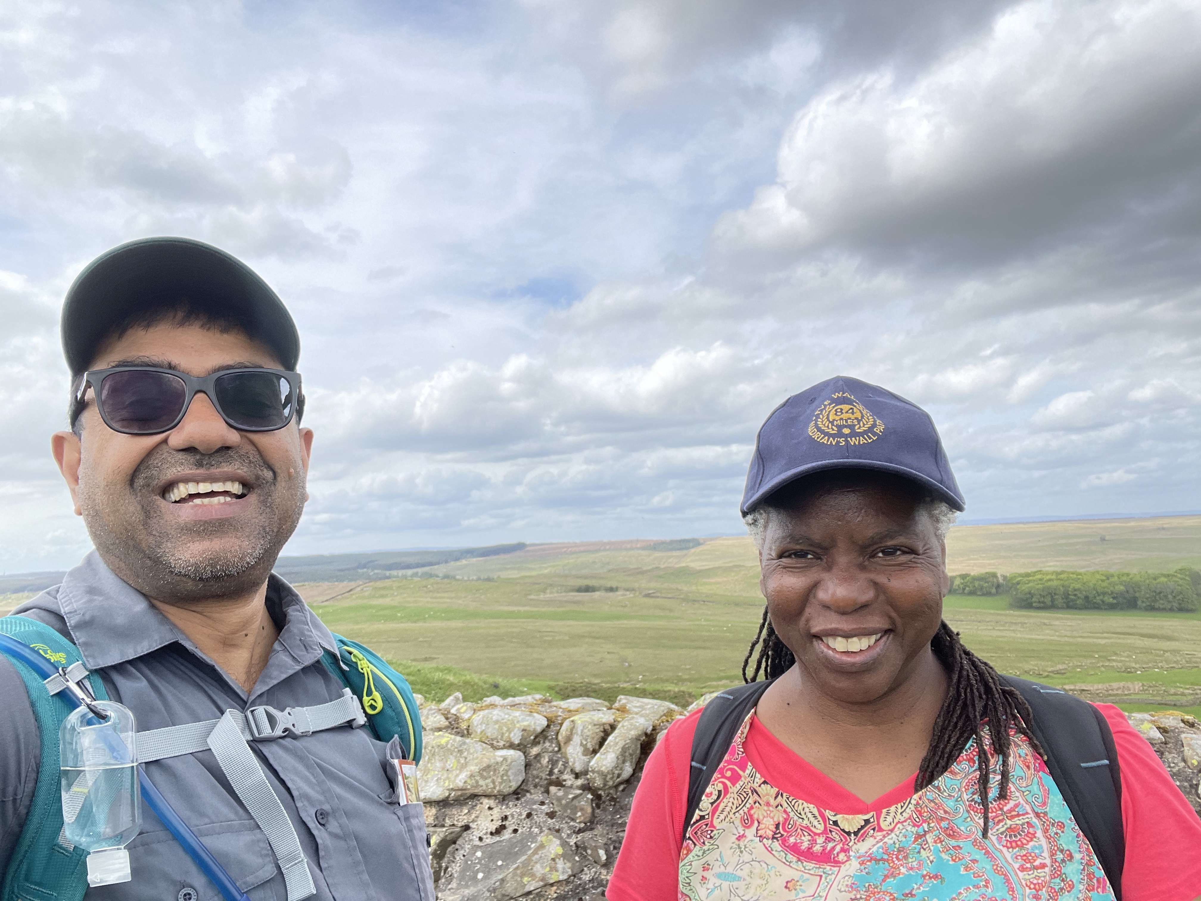 Blog author, Mohammed Dhalech and a female colleague, both wearing caps stand in front of Hadrian's Wall Path smiling at the cameraing caps