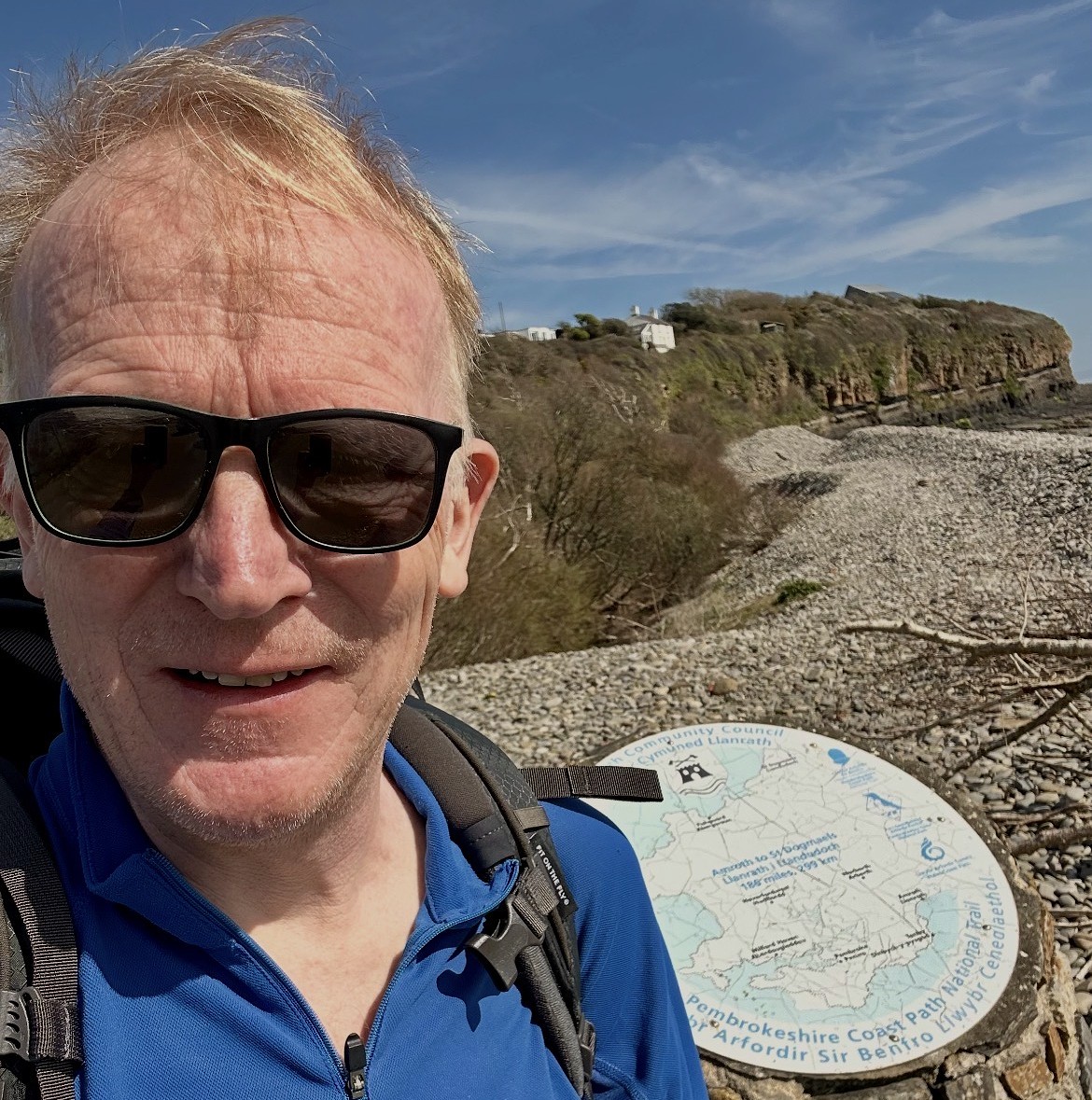 The blog Author Paul Gamble who lives with AuDHD takes a selfie at the end of the Pembrokeshire Coast Path. It's a sunny day and he is wearing dark glasses. In the background is a plaque marking the end of the National Trail and beyond that the Pembrokeshire coastline stretches into the distance.