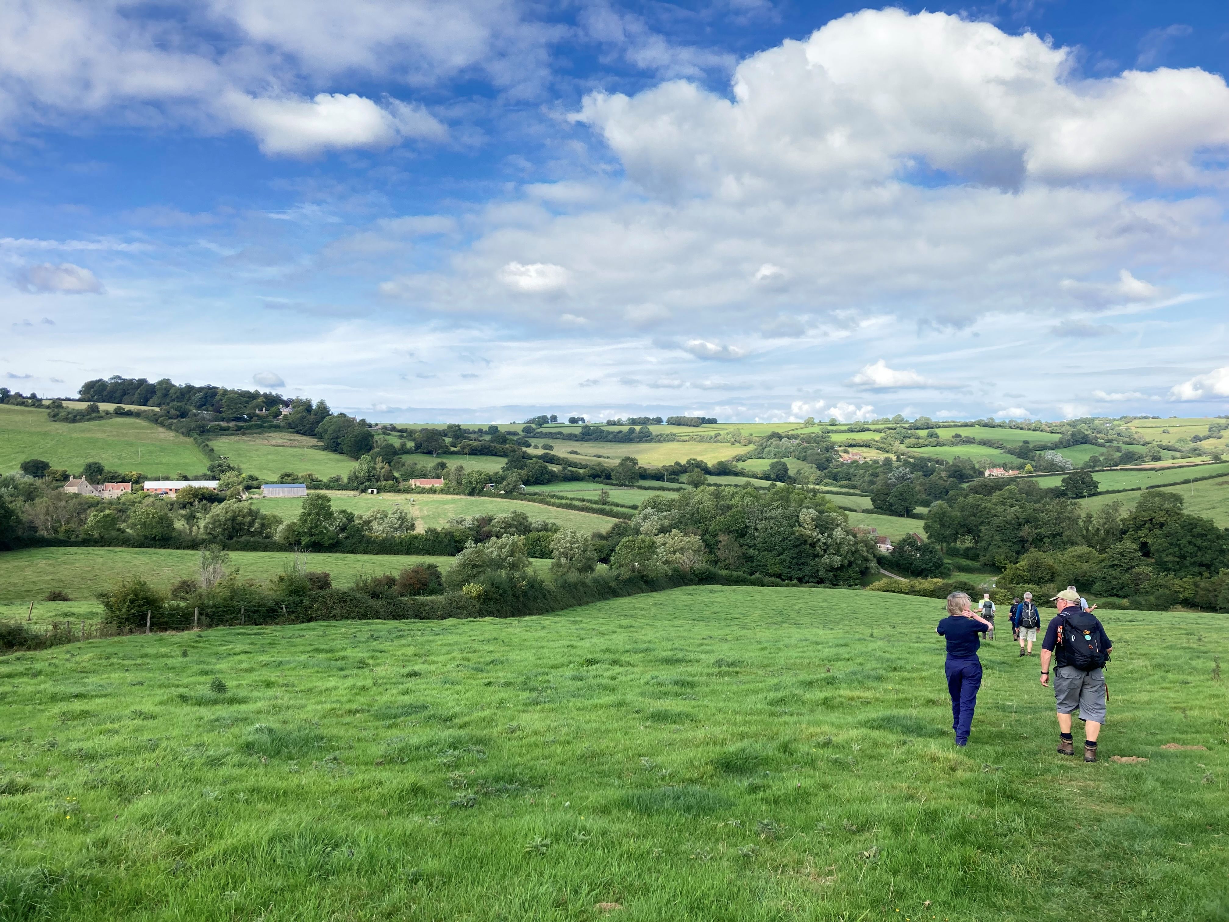 A group of people walk along the Cotswold Way on a sunny day with clouds scudding across a blue sky