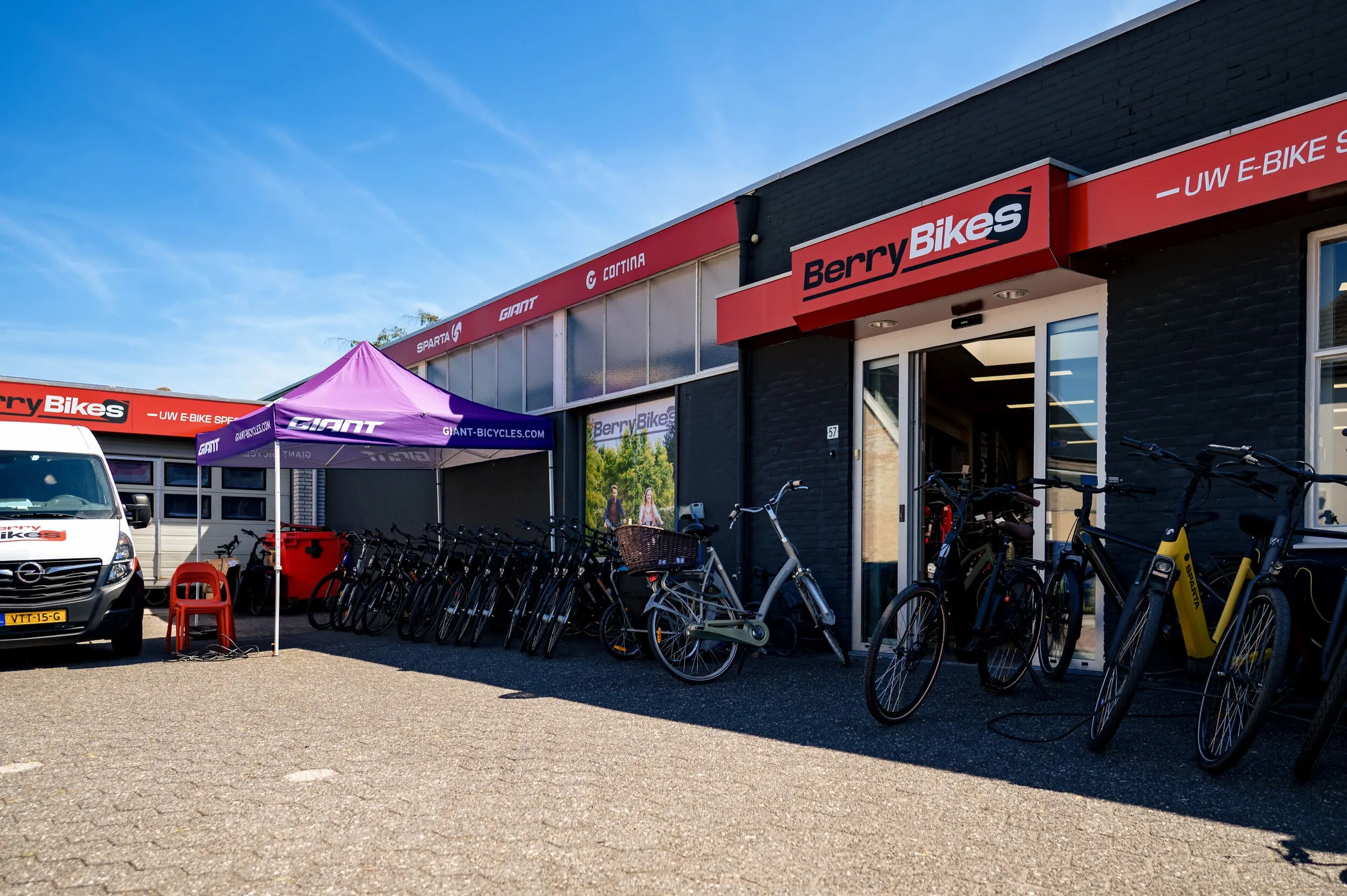 Fietsenwinkel Berry Bikes met een rij fietsen buiten, een bestelwagen en een paarse Giant-tent onder een blauwe lucht.