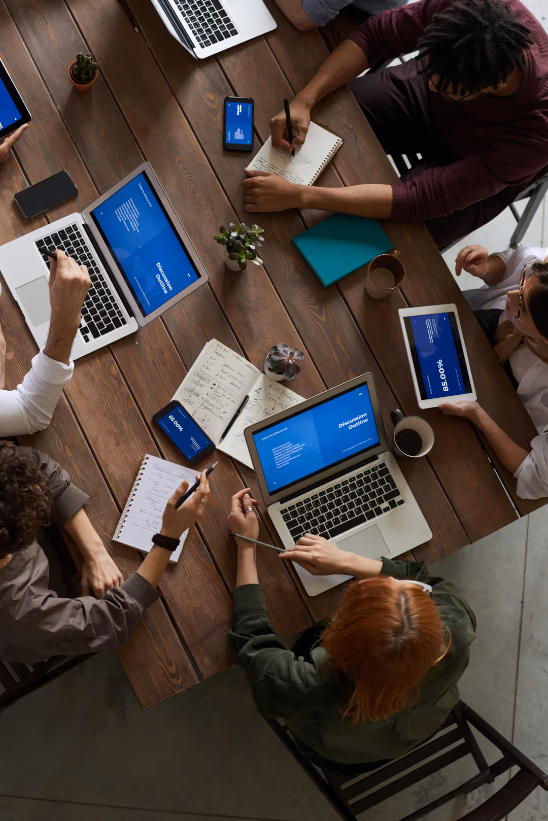 Overhead view of a group of people collaborating around a wooden table with laptops, tablets, smartphones, notebooks, and coffee cups.