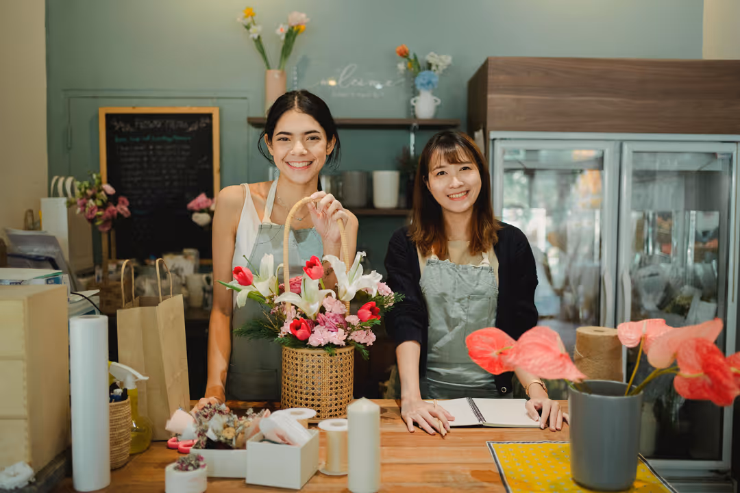 Two smiling women wearing aprons standing behind a wooden counter with a flower arrangement and notebooks in a flower shop.