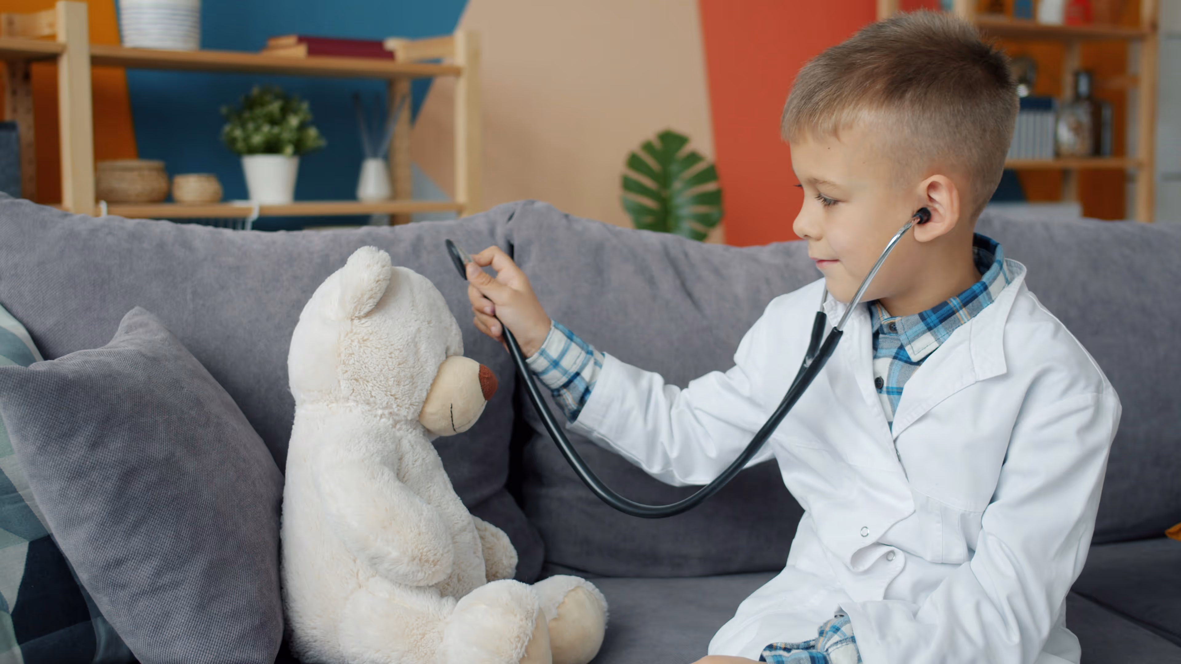 Young boy wearing a white coat playing doctor with a stethoscope on a teddy bear seated on a couch.