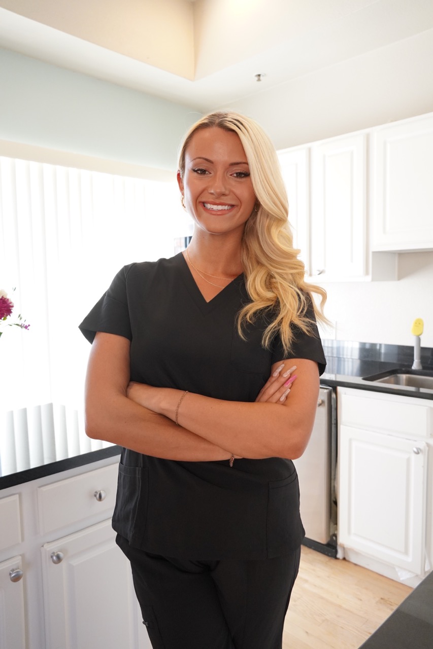 Smiling woman with long blonde hair wearing black scrubs standing with arms crossed in a bright kitchen.