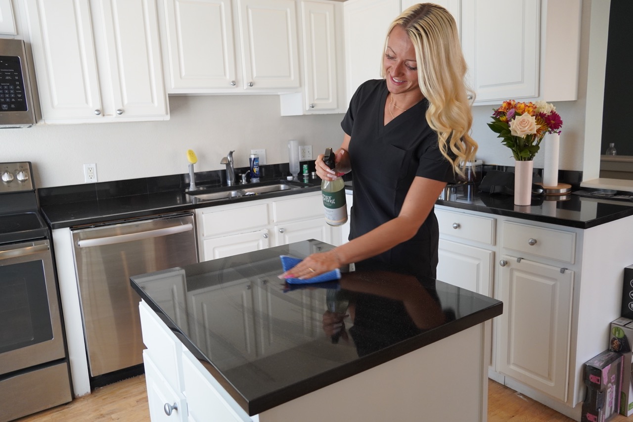 Smiling woman with long blonde hair cleaning a black kitchen island countertop with a spray bottle and blue cloth in a modern kitchen.