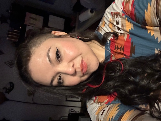 Woman with long curly dark hair with pink highlights, wearing colorful patterned clothing and eyeliner, posing indoors.