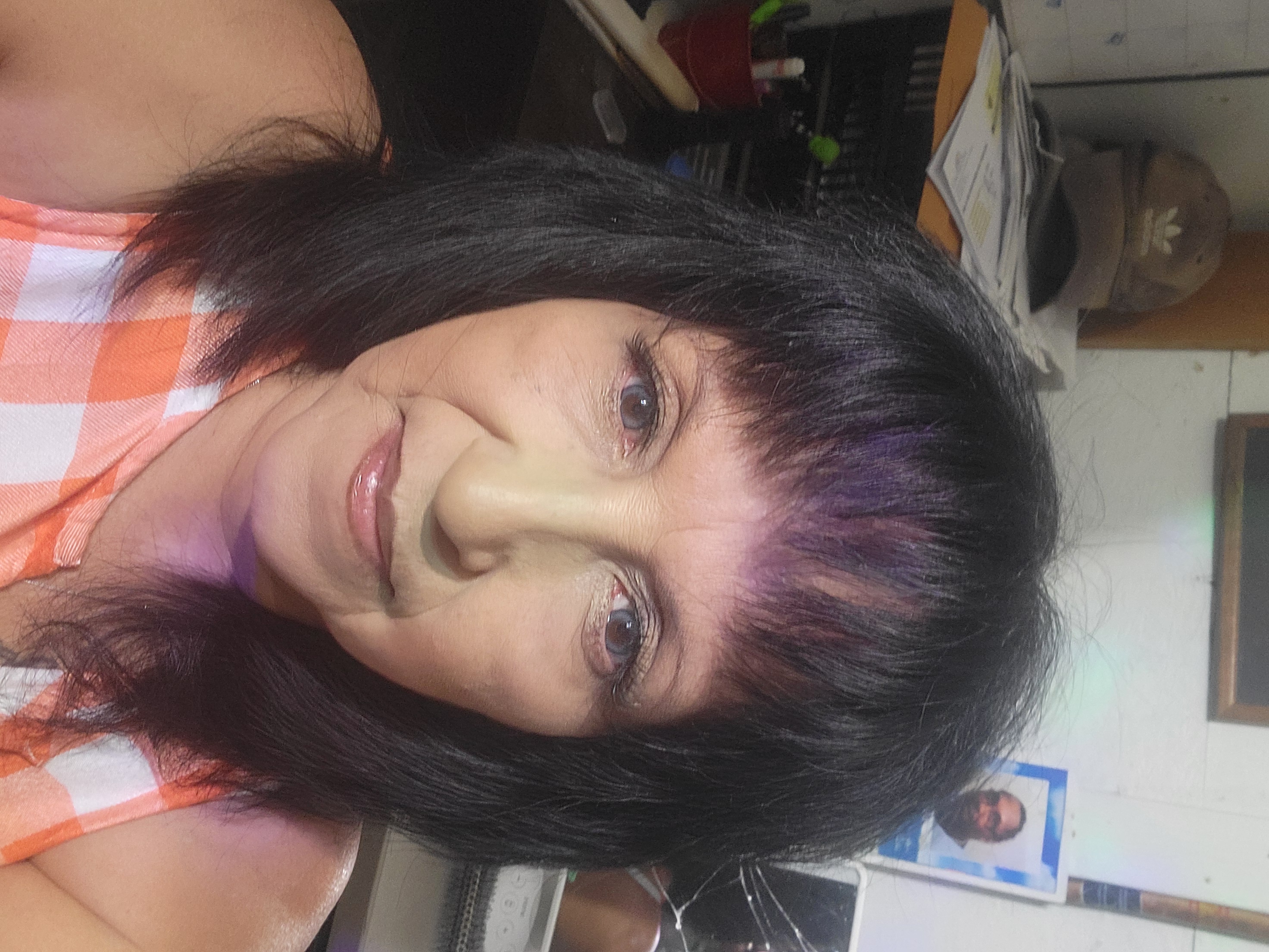 Middle-aged woman with black hair and bangs wearing an orange and white checkered top, looking at the camera indoors.
