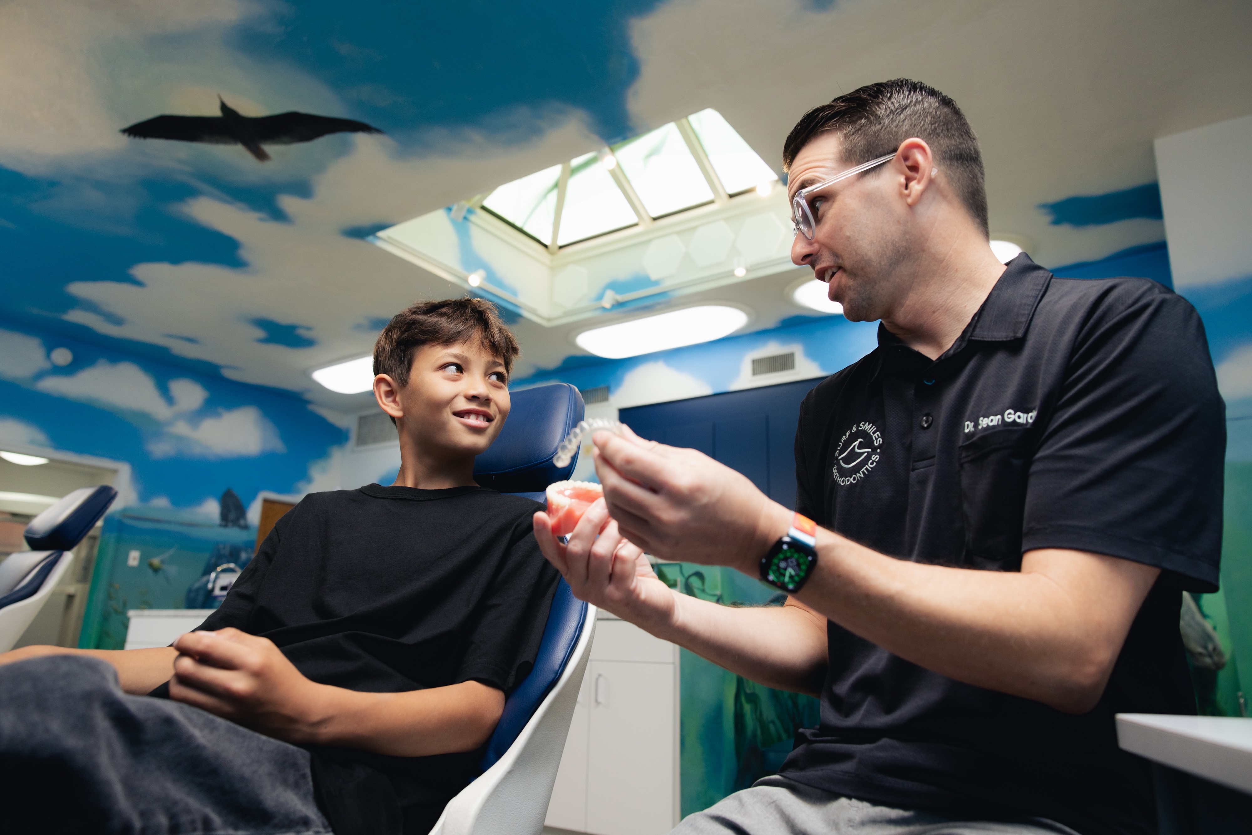 The orthodontist is explaining how Invisalign works to a young boy who is sitting in the dental office chair.