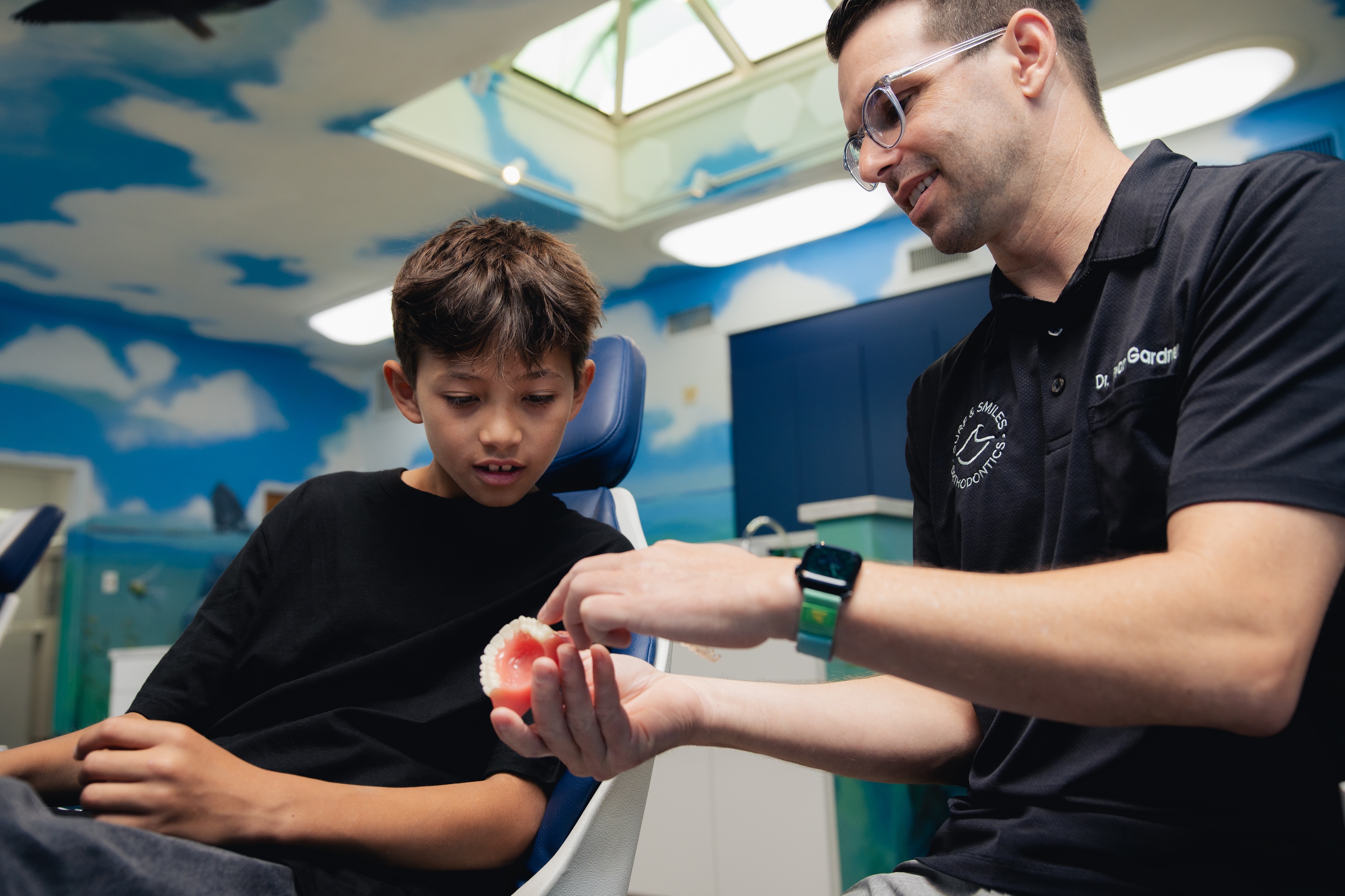 The orthodontist is showing a young boy a 3D model of a set of teeth and explaining how braces work.