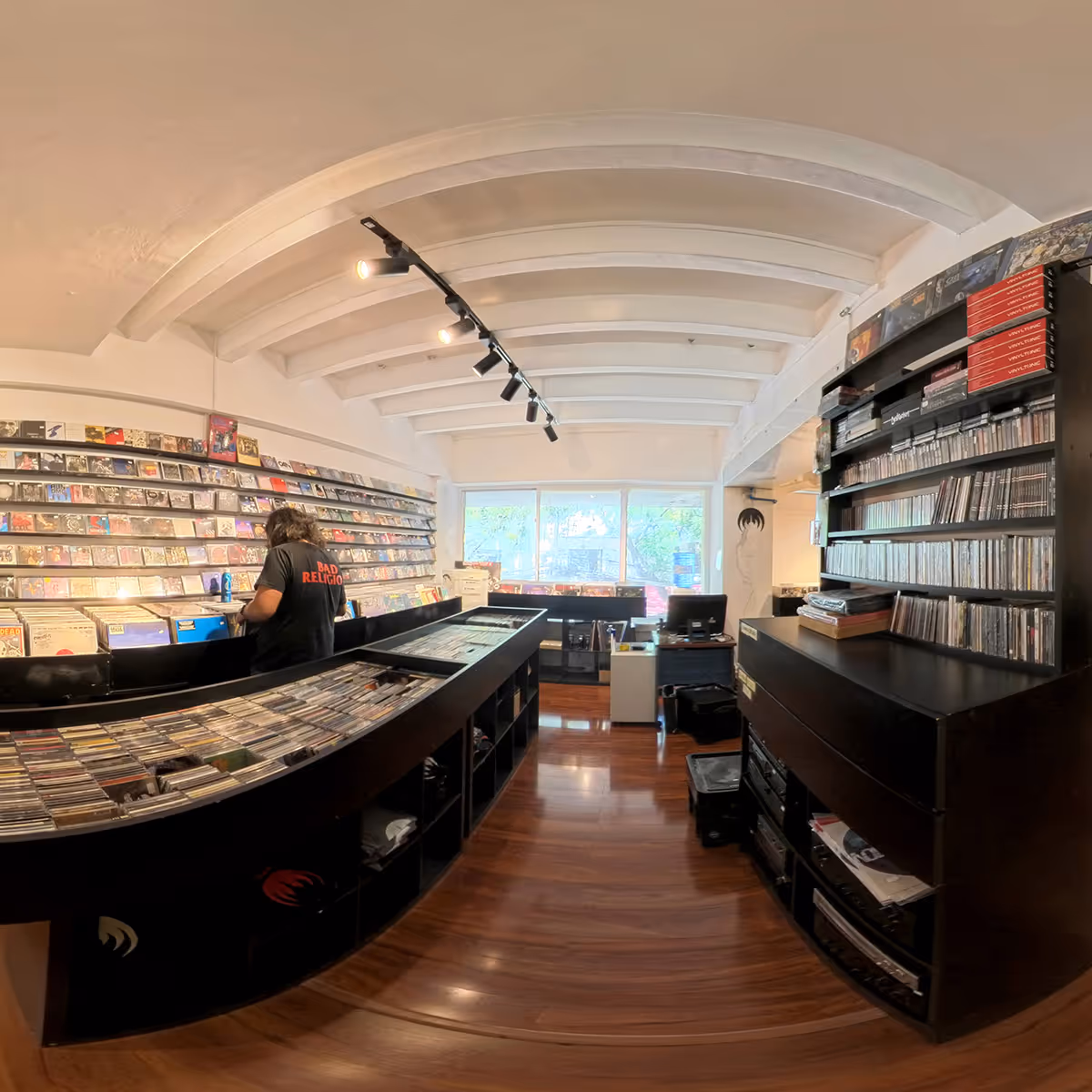 Interior of a record store with wooden floors, shelves filled with CDs and vinyl records, and a person browsing.