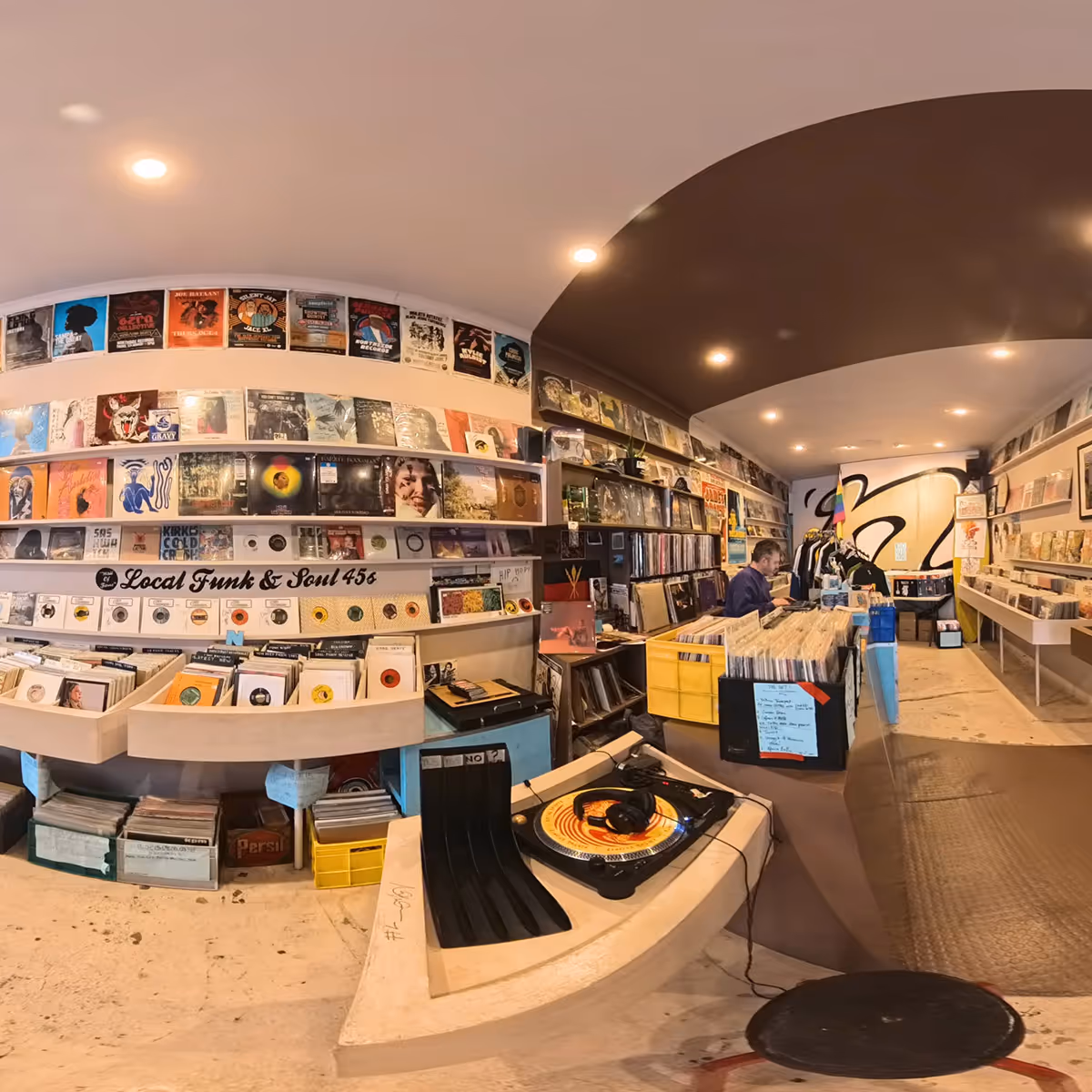 Interior of a cozy record store with vinyl records displayed on shelves, a turntable in the foreground, and a person working behind the counter.