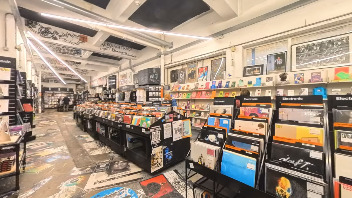 Virtual tour interior view of Rough Trade East record store in London, showing vinyl record displays, browsing customers, and the shop's distinctive industrial warehouse aesthetic with exposed ceiling beams and concrete floors