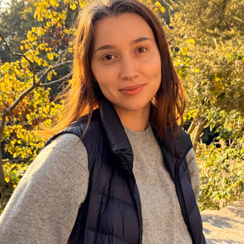 Young woman wearing a black vest and gray sweater standing outdoors with trees in autumn light.