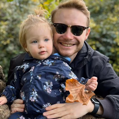 Smiling man wearing sunglasses holds a toddler in a floral jacket holding a large brown leaf outdoors.