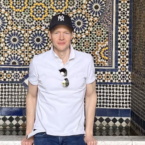 Man wearing a white polo shirt and black NY baseball cap standing against a colorful geometric tiled wall.