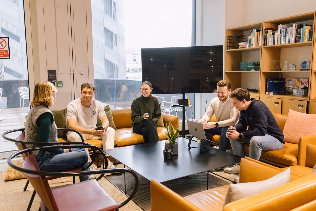 Five people sitting and chatting in a cozy office lounge with couches, a TV, and bookshelves.