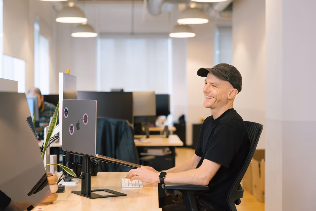 A man in a black cap and shirt smiles while sitting at a desk with a laptop in a modern office.