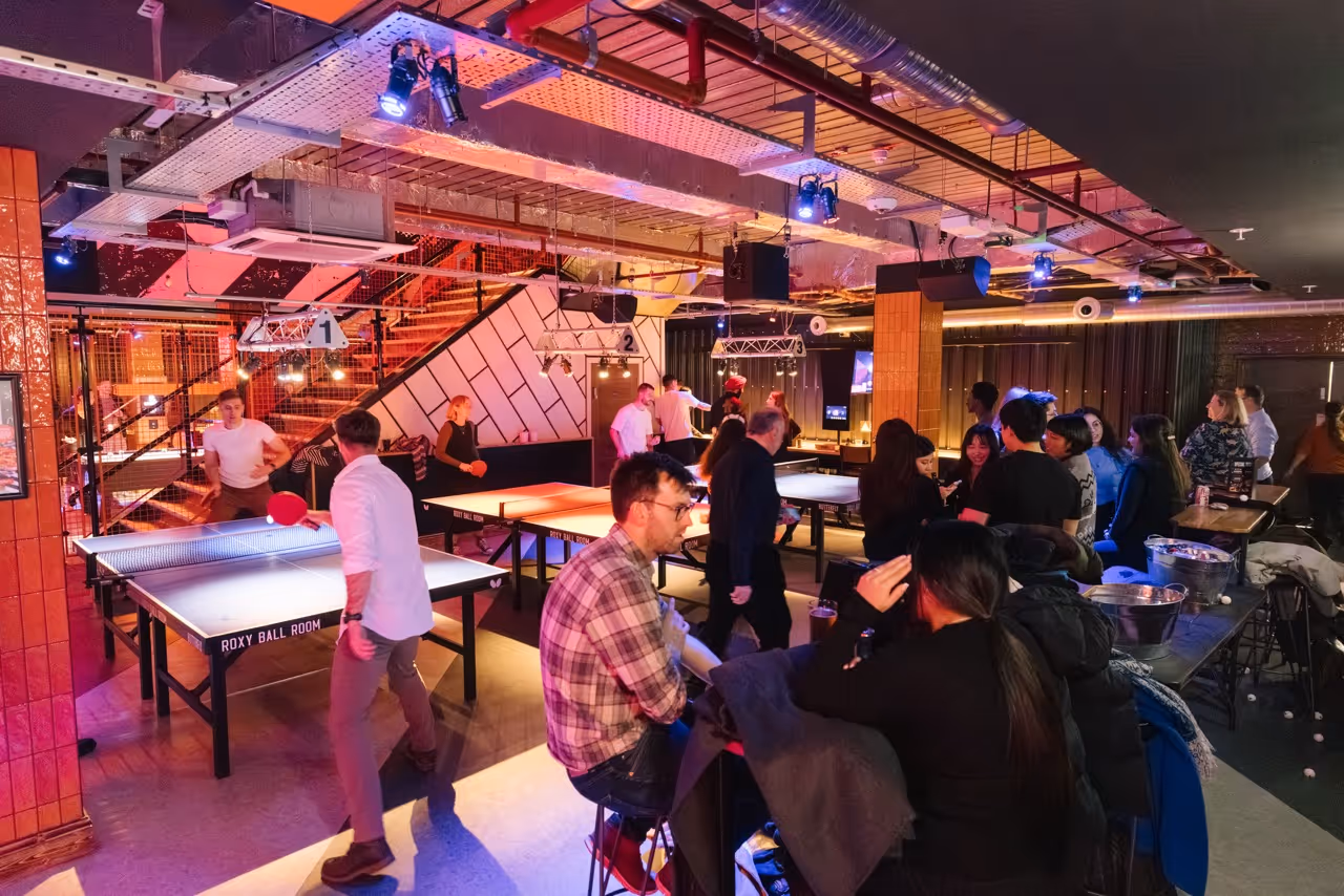 People playing and watching table tennis in a lively game room with modern lighting and decor.