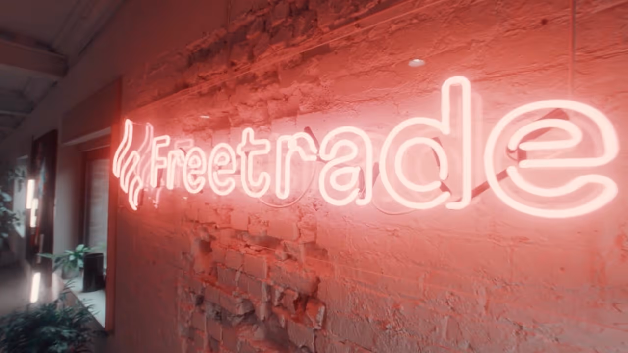 Pink neon sign spelling 'Freetrade' mounted on a textured brick wall in an indoor space.