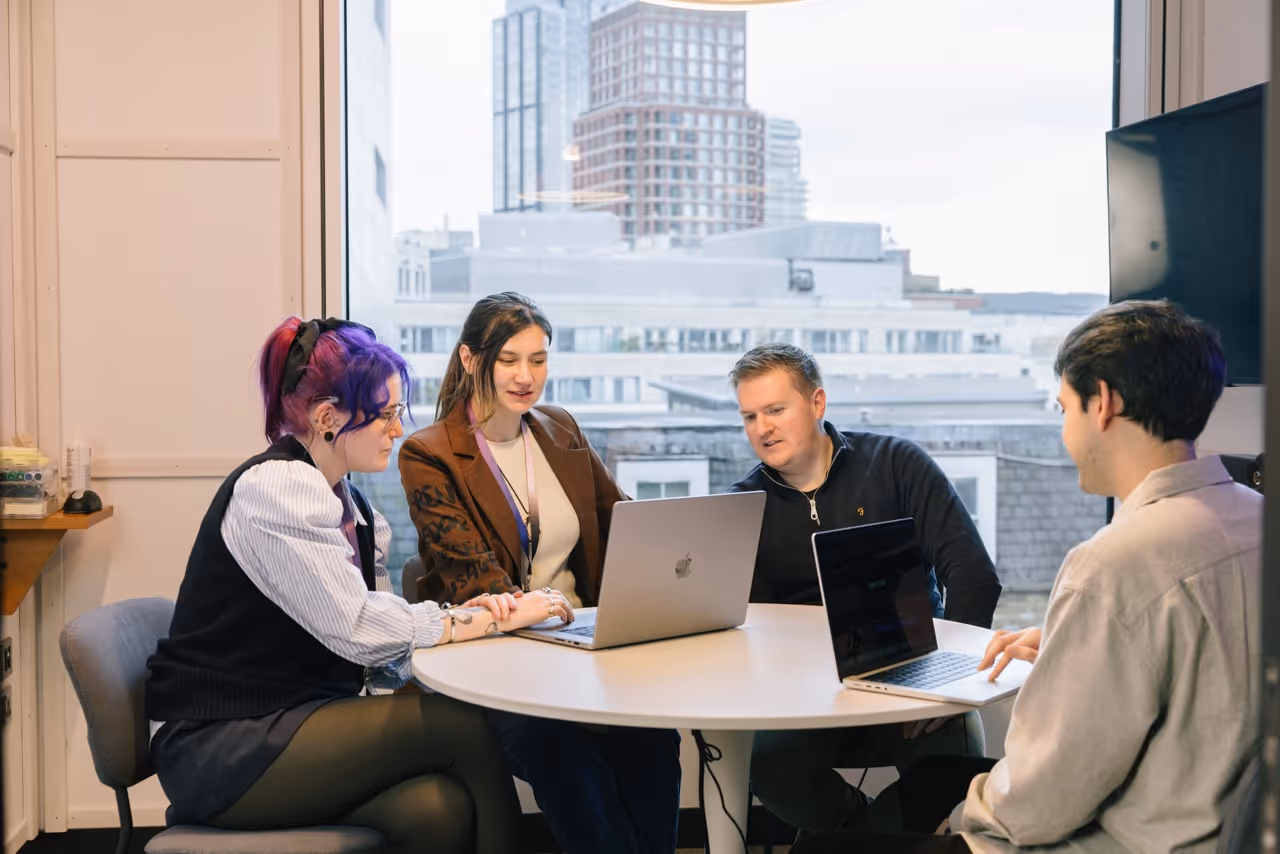 Four people sitting around a round table working on laptops in an office with city view windows.