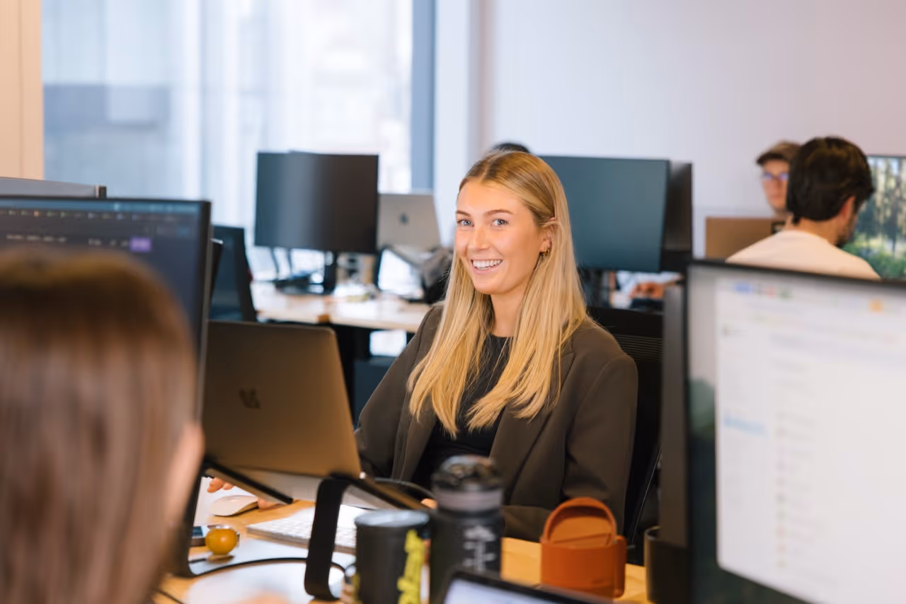 Smiling woman with long blonde hair sitting at a desk with a laptop in a modern office.