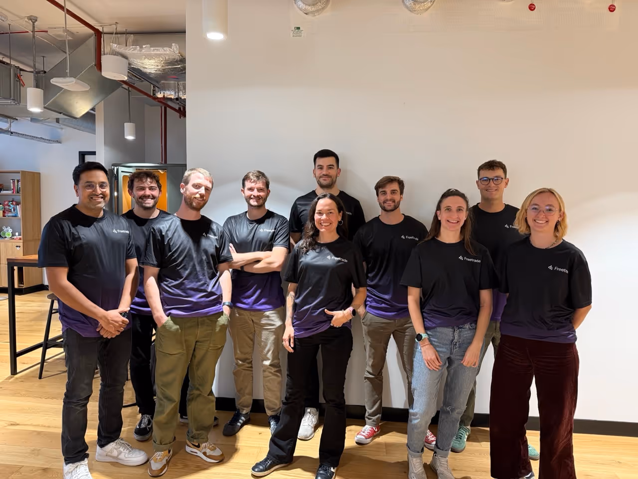 Group of ten smiling people wearing black and purple Freetrade T-shirts standing indoors.
