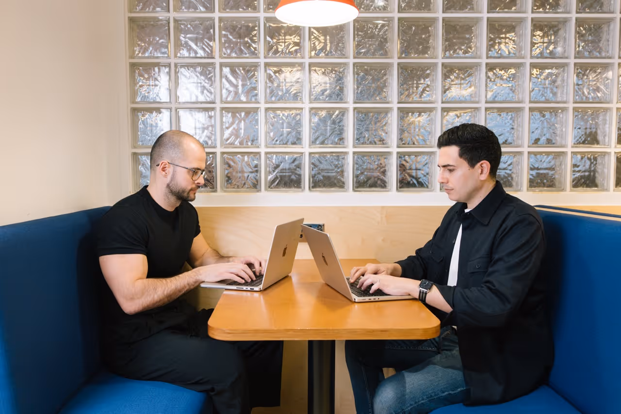 Two men sitting across from each other at a wooden table working on Apple laptops in a modern office.