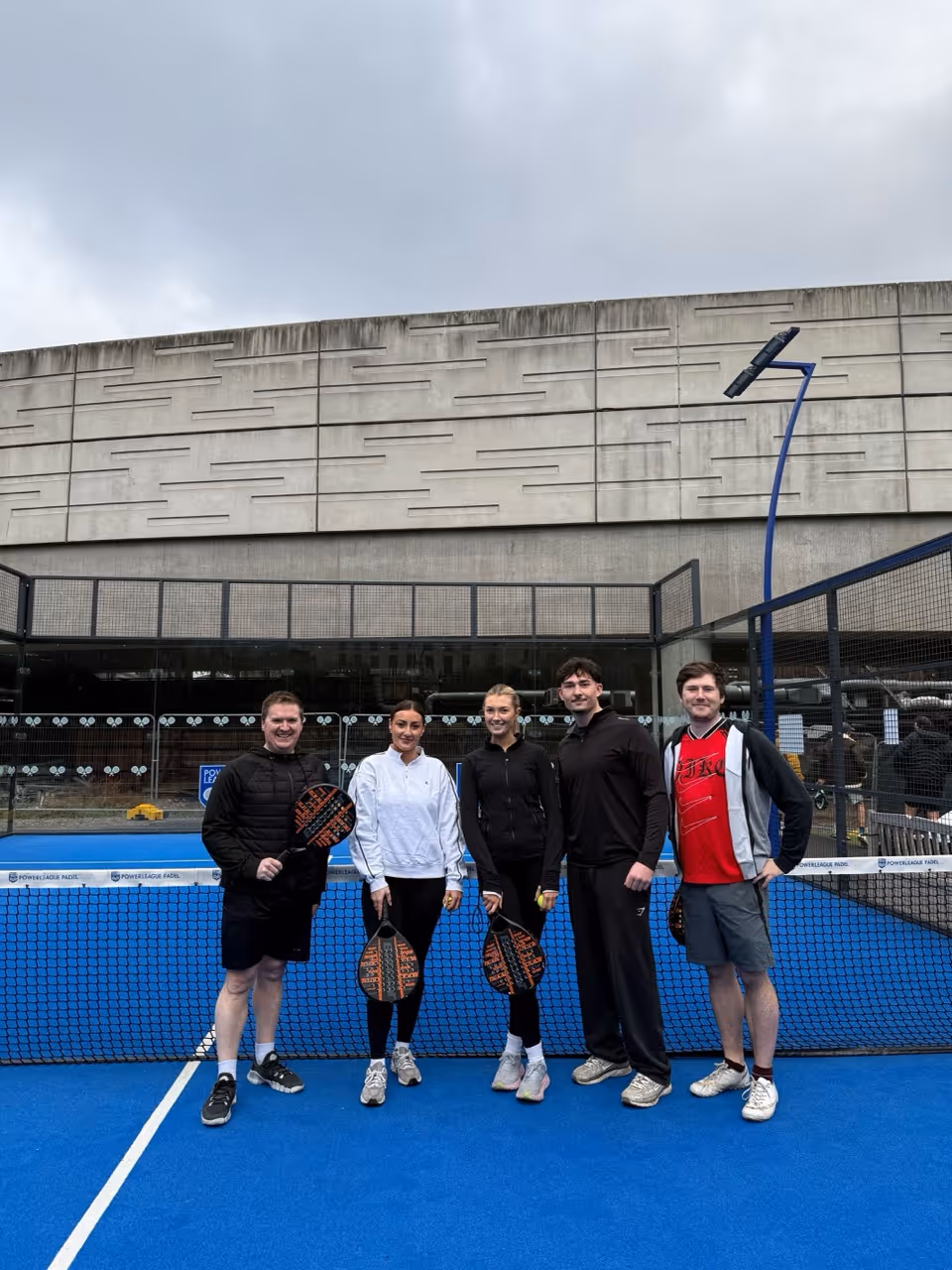 Five people posing on a blue padel court holding paddles, with a concrete building background.