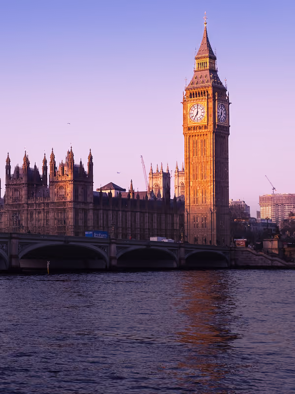 Sunlit Big Ben clock tower and part of the Palace of Westminster beside the River Thames in London during a clear evening with a purple sky.