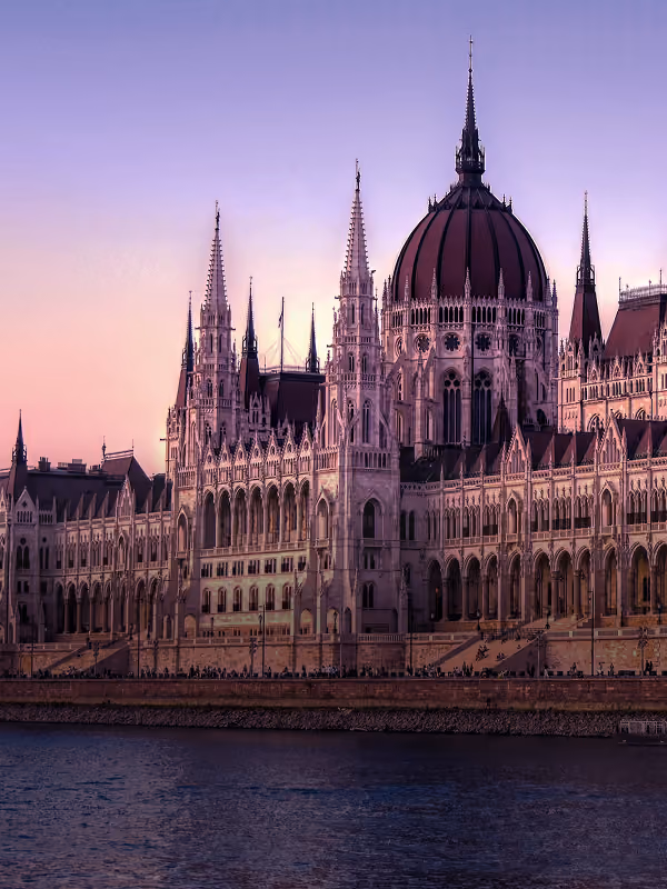 The Hungarian Parliament Building at dusk with its Gothic Revival architecture illuminated under a purple-pink sky beside the Danube River in Budapest.