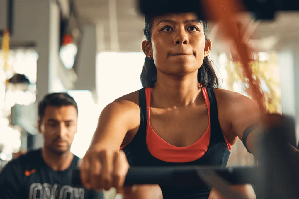 Focused woman rowing on a machine in a gym with a trainer watching behind her.