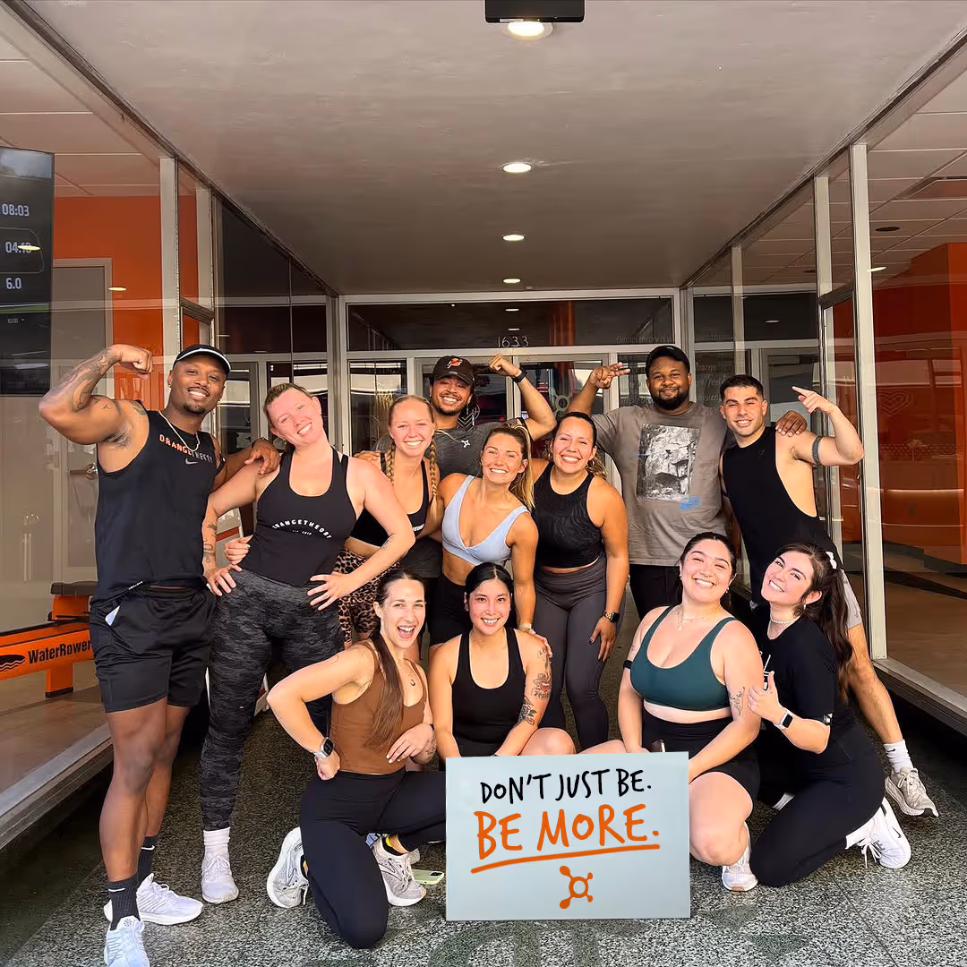 Group of smiling diverse people in athletic wear posing indoors, holding a sign that says 'DON'T JUST BE. BE MORE.'