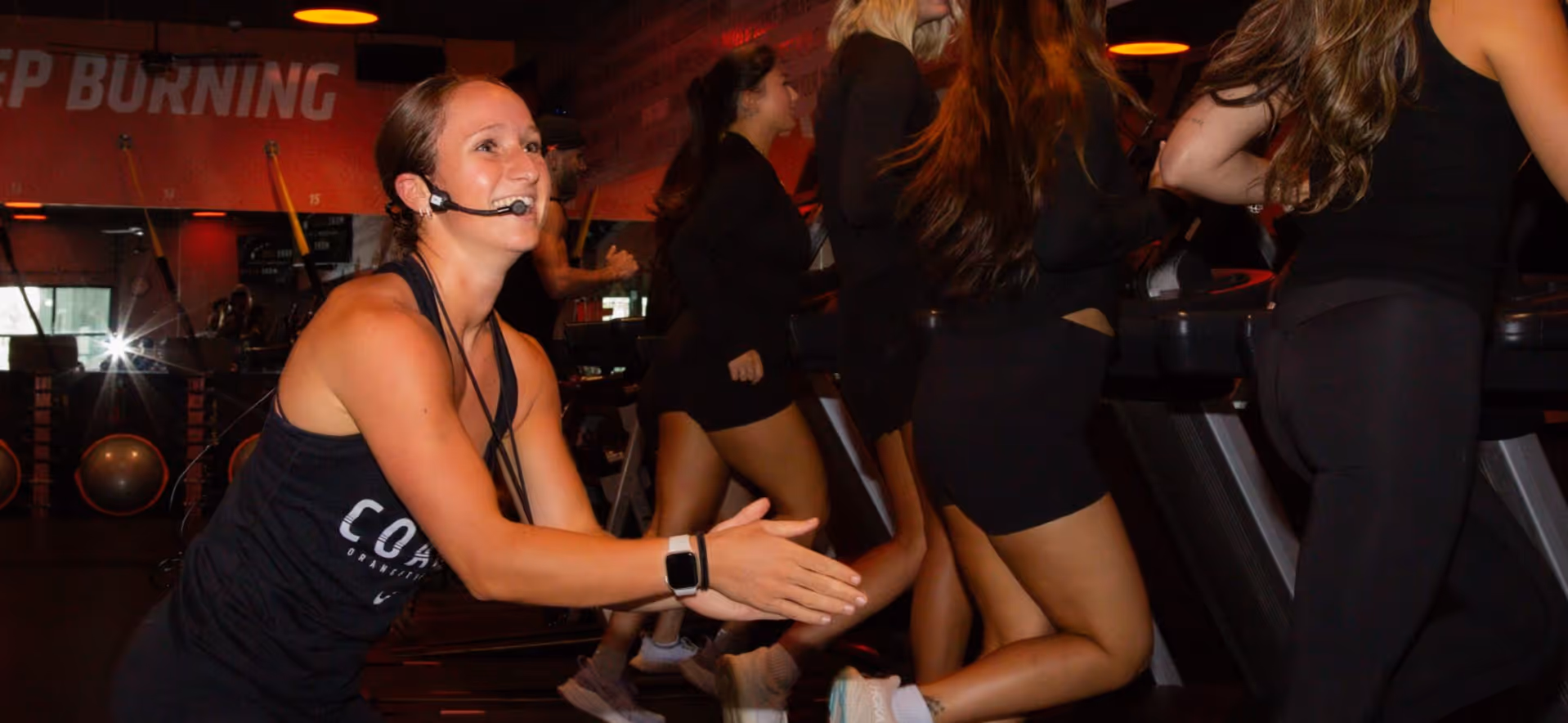 Fitness coach enthusiastically leads a group workout class as participants run on treadmills in studio.