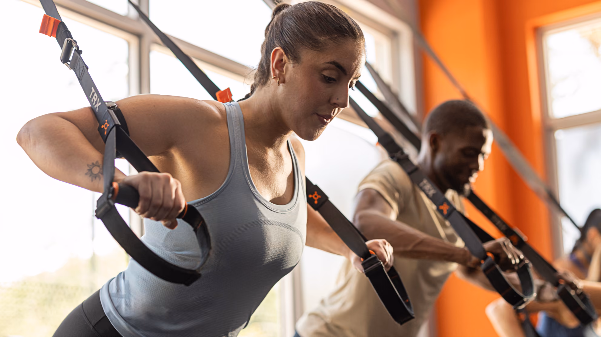 Focused man and woman using TRX straps for strength training in a bright gym with orange walls.