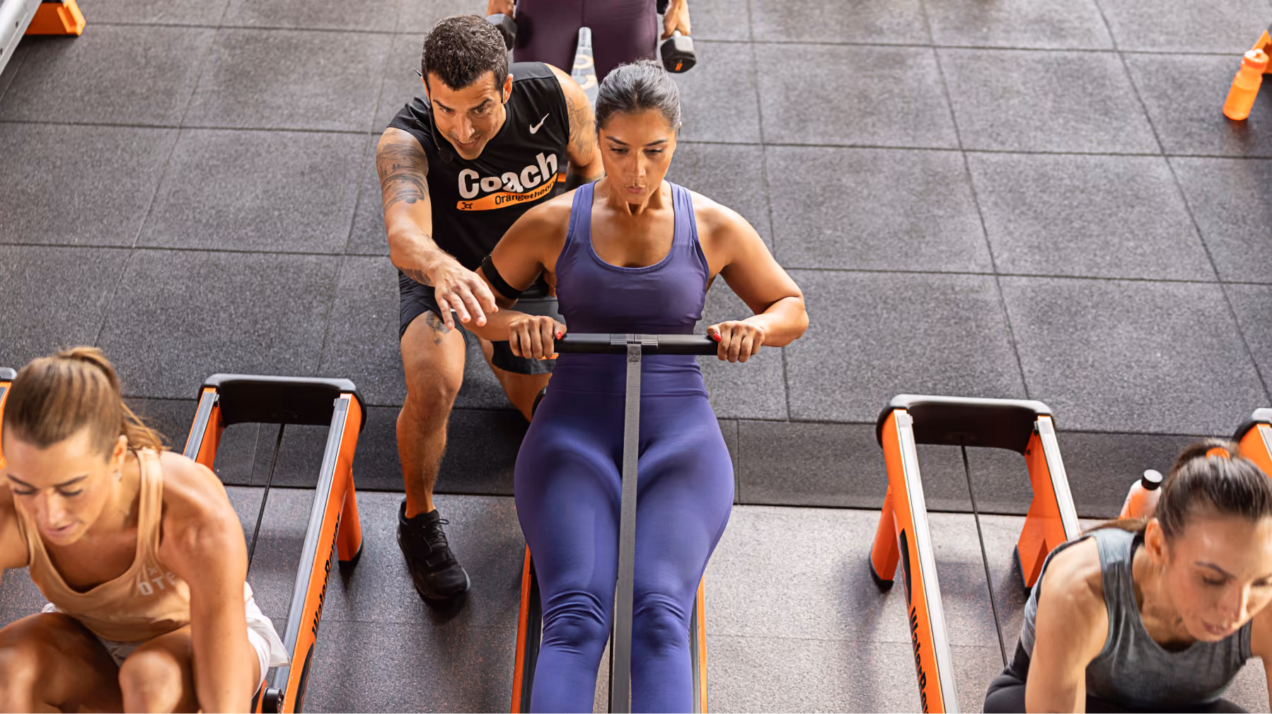 Woman on rowing machine coached by trainer in gym with others rowing on similar machines.