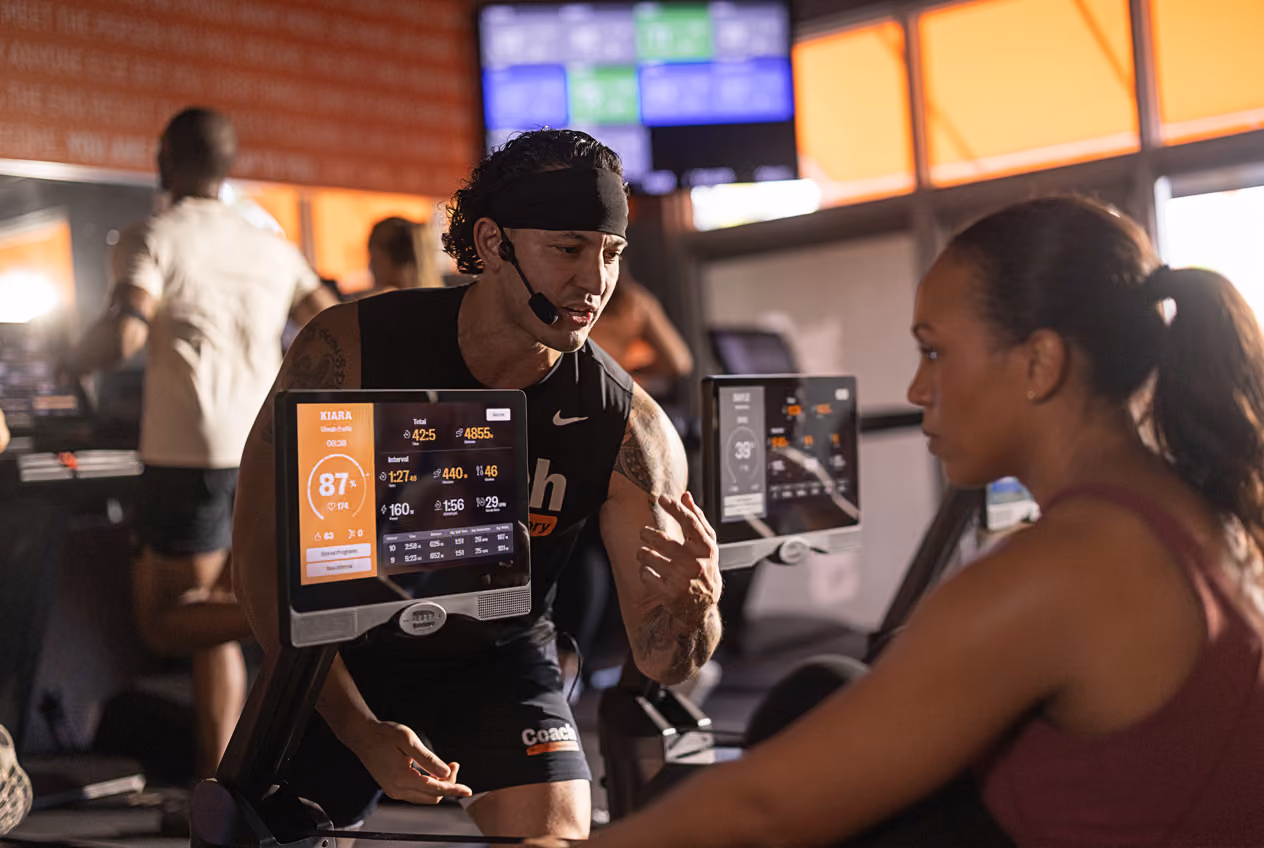 Fitness coach wearing headset instructs woman working out on rowing machine at gym.