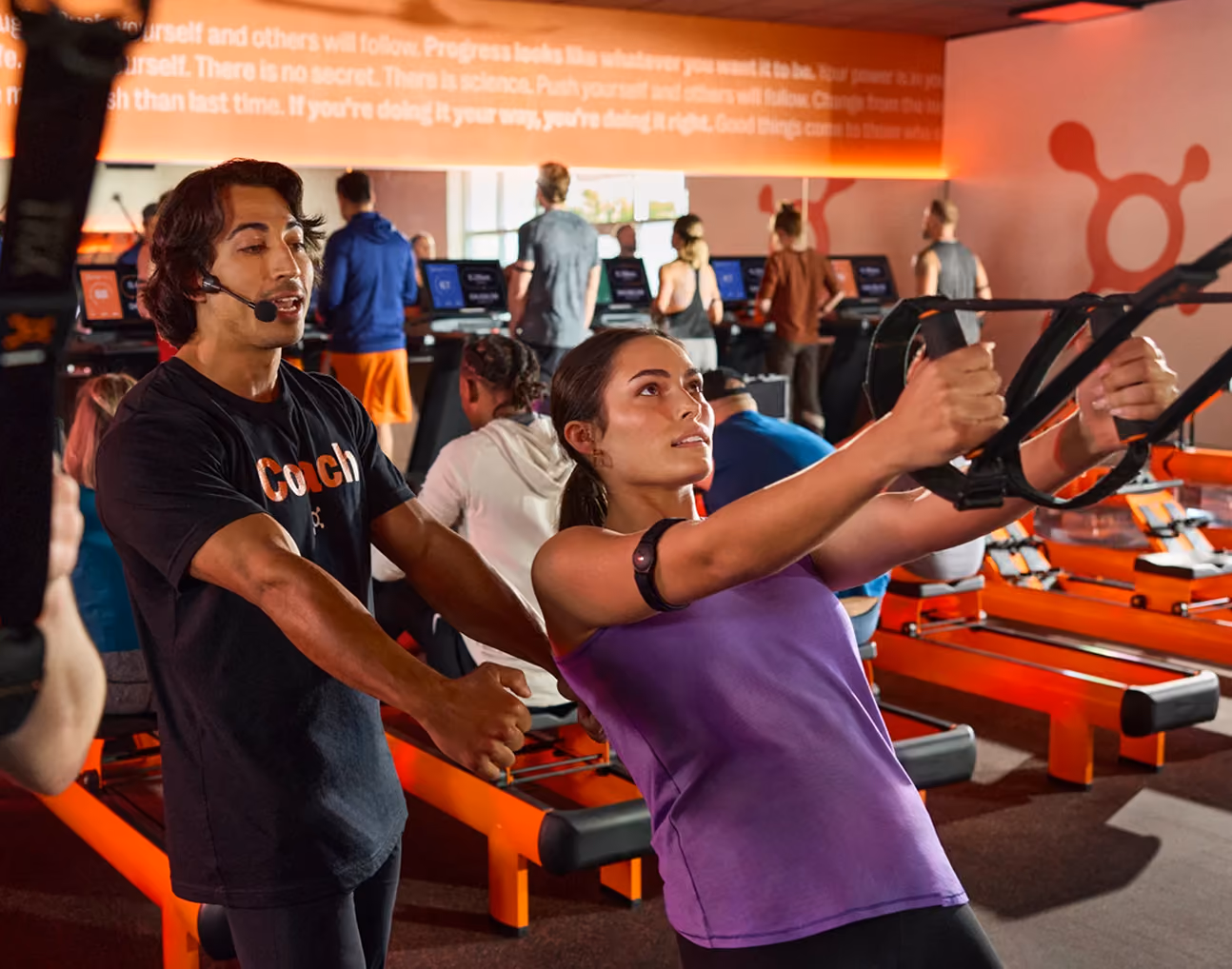 A female athlete in a purple tank top exercises with resistance straps while a male coach in a black shirt and headset assists her in a gym with treadmills and other people working out in the background.