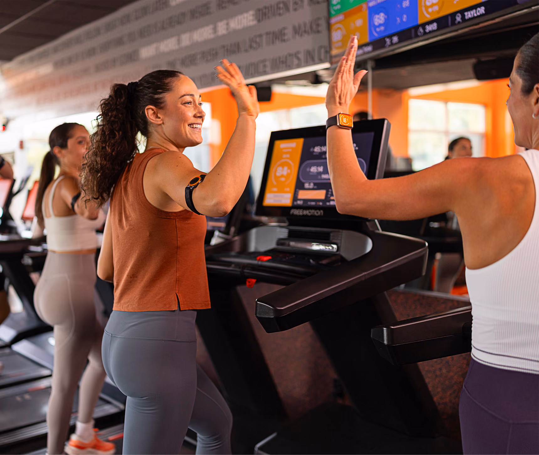 Dos mujeres en máquinas de correr en un gimnasio chocando las manos, una viste una blusa naranja sin mangas y la otra una blusa blanca sin mangas, ambas sonriendo durante el ejercicio glosario.