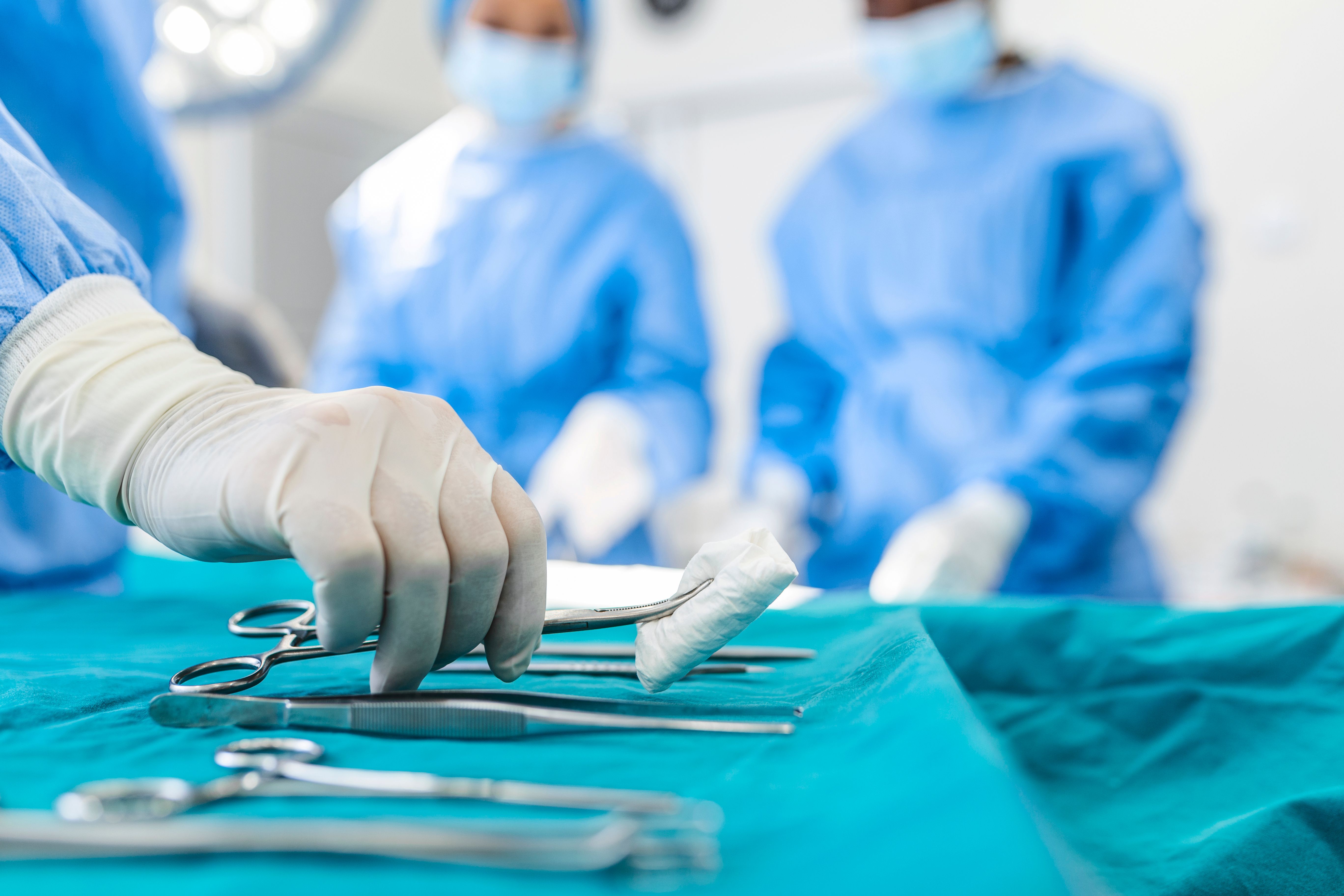 Three unidentifiable medics in a hospital surgery room with one person picking up medical implements