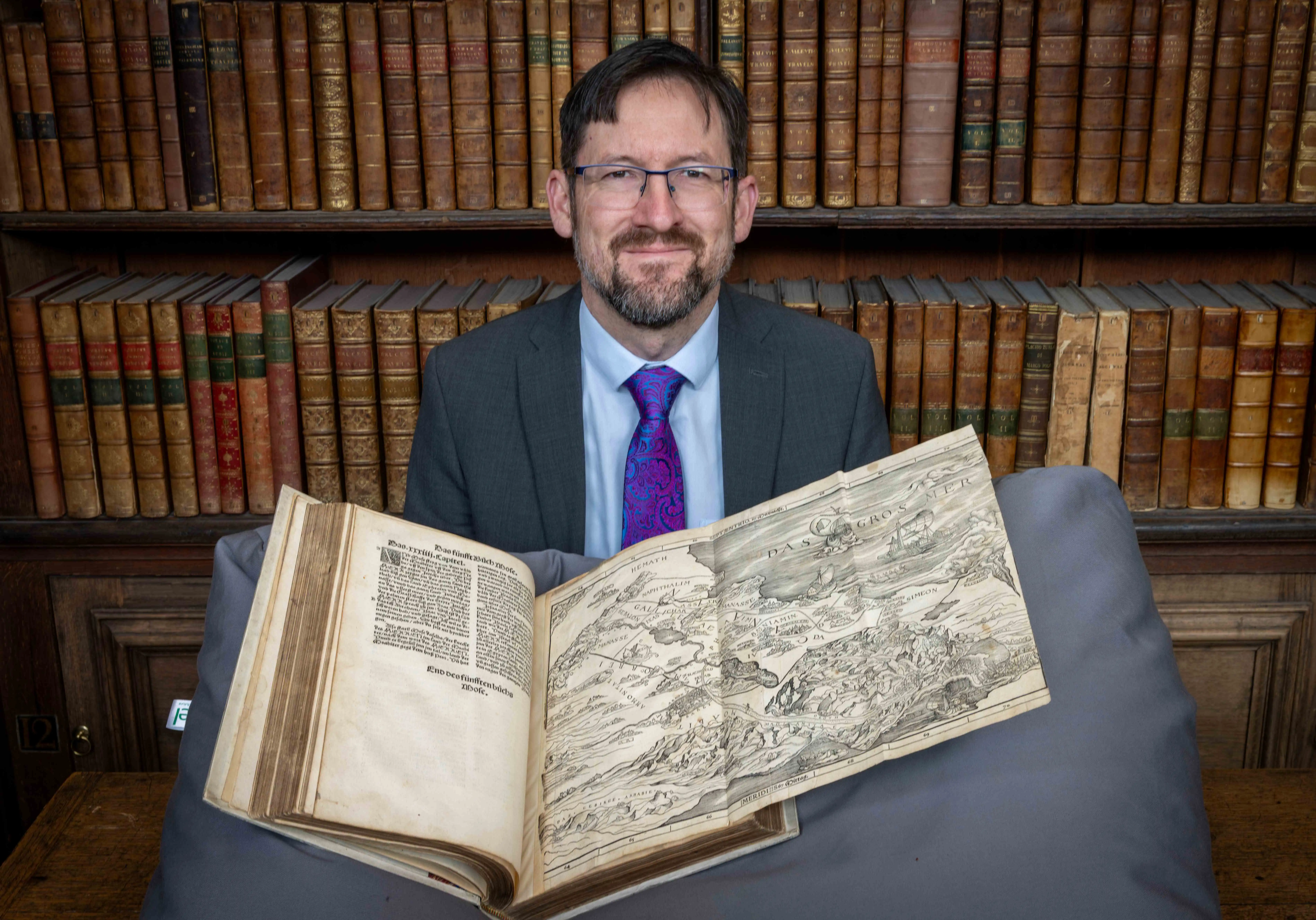 Professor Nathan MacDonald holds open the page of the Bible showing the map with shelves of antique books behind him