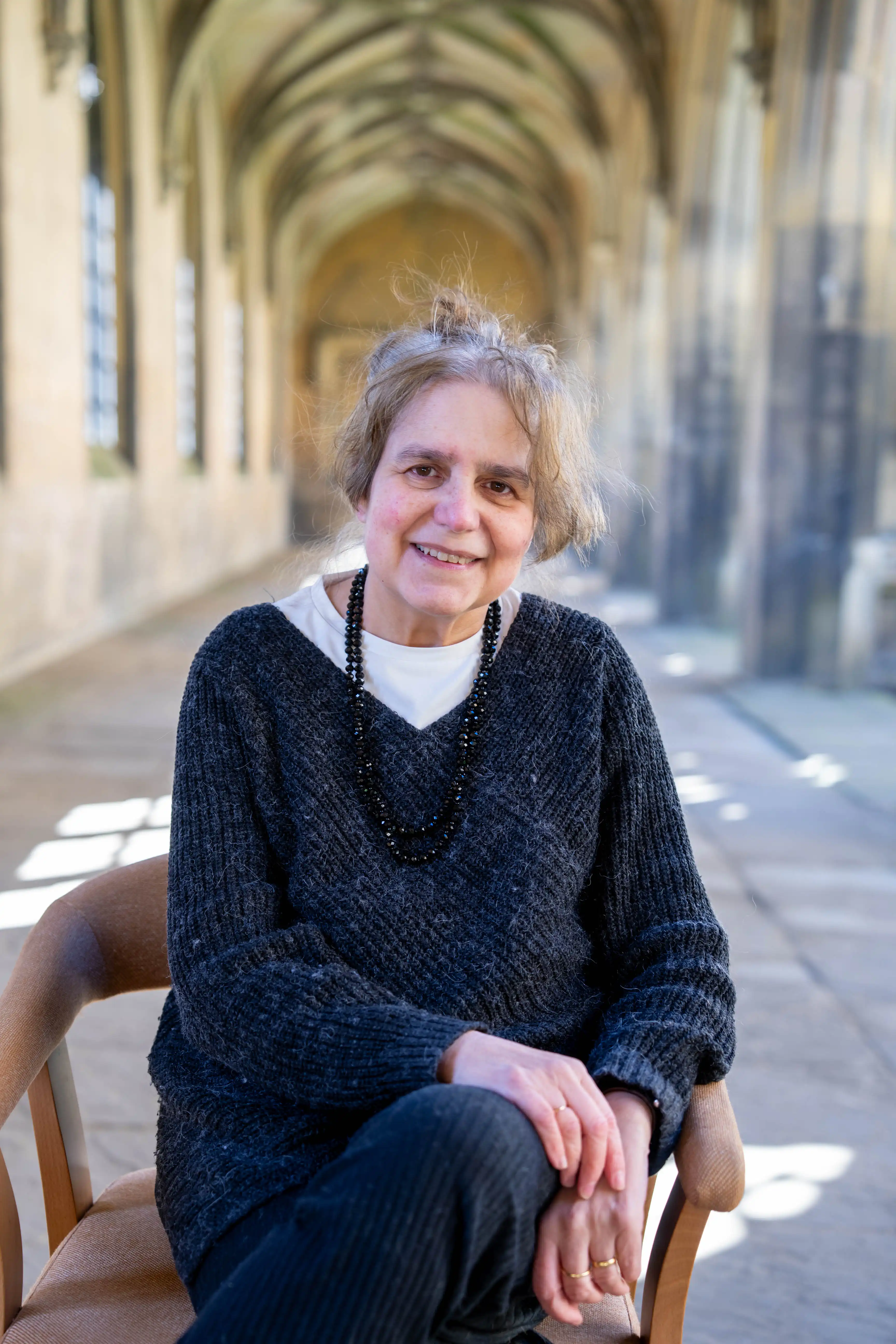 A portrait photo of Professor Maria Manuel (Manucha) Lisboa sitting on a chair in The Cloisters, New Court, St John's College