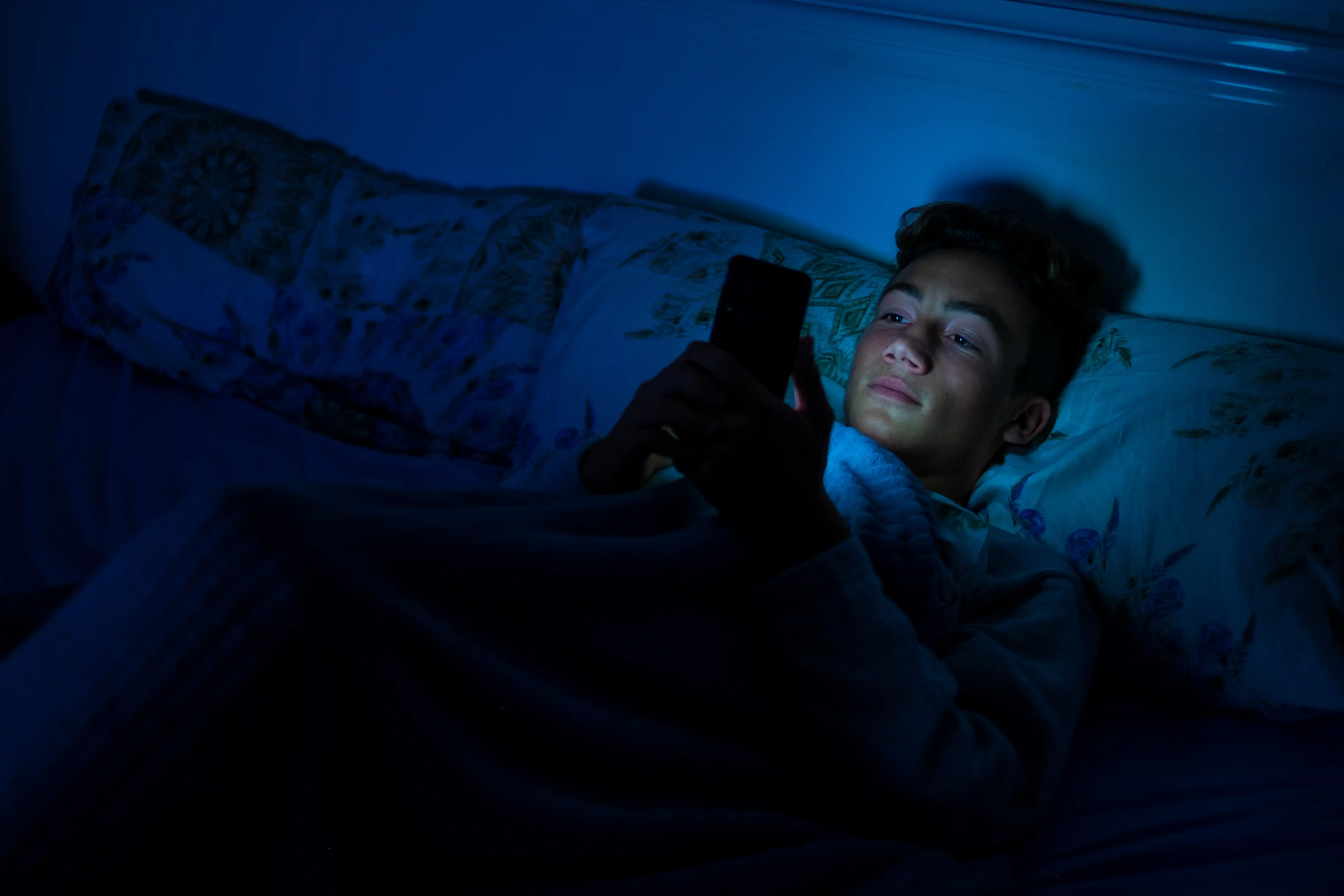 Teenage boy lays in bed in the dark looking at a smartphone with his face lit up by the light emitting from it