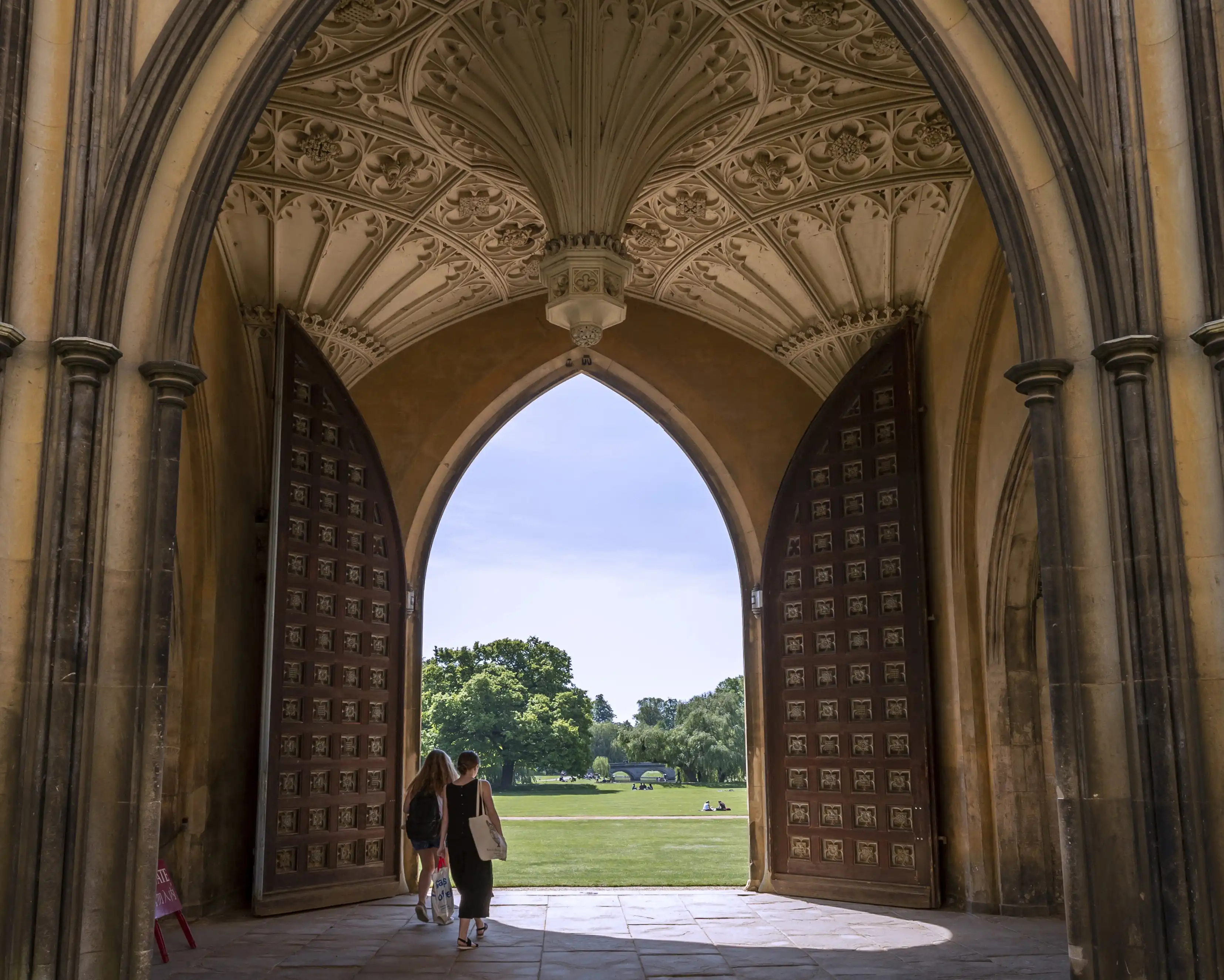 Two people walk through Eagle Gate, New Court, St John's College on to the College Backs in summer time
