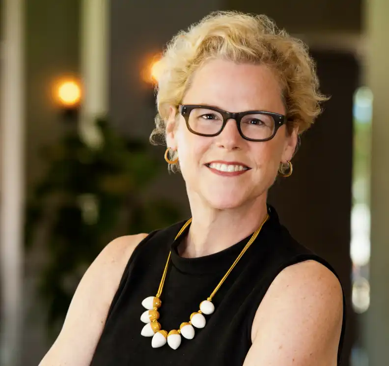 Joy Connolly is wearing a sleeveless black top and a beaded necklace, standing with arms crossed indoors.