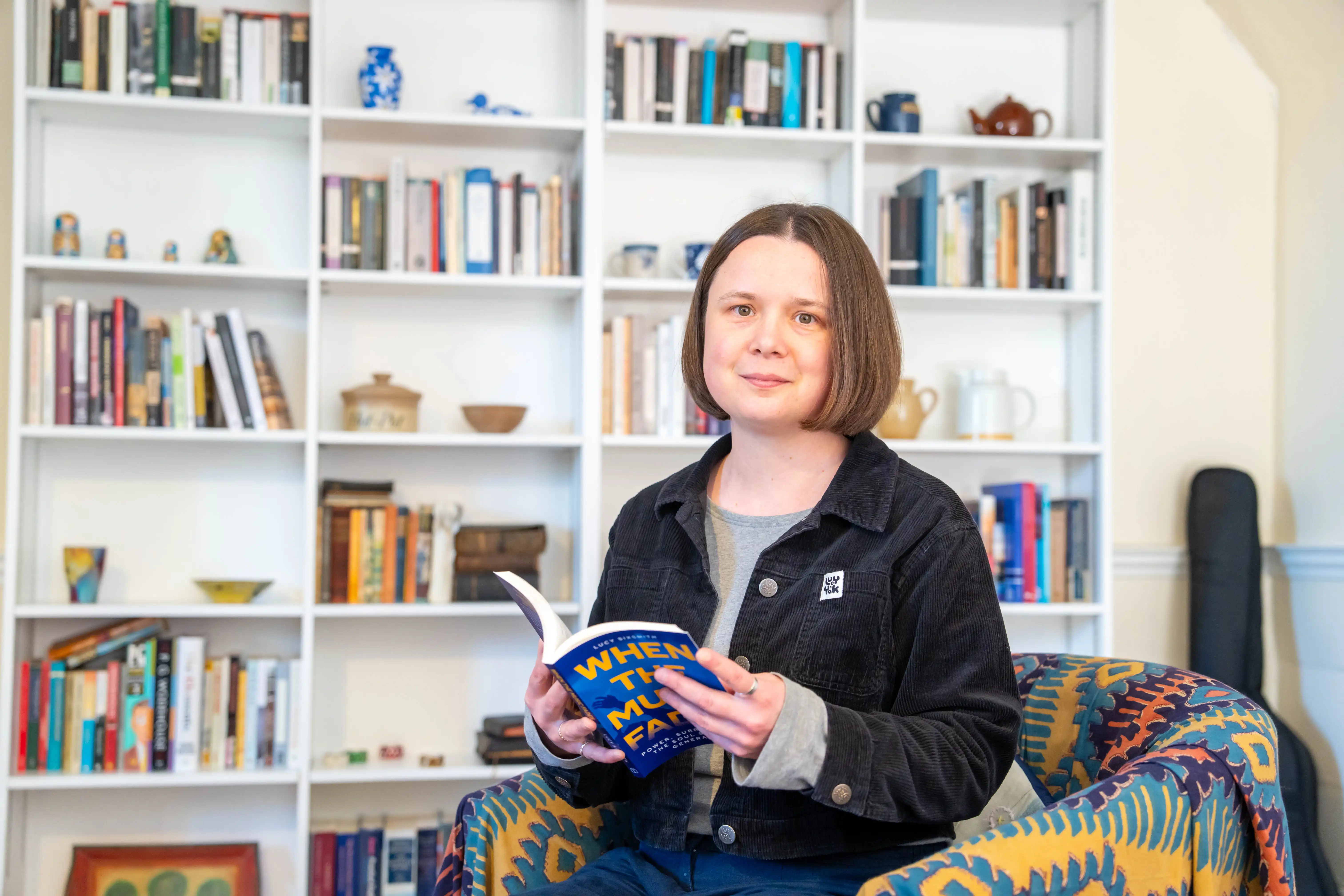 Dr Lucy Sixsmith sits in her study with her book open in her hands, with bookshelves behind her